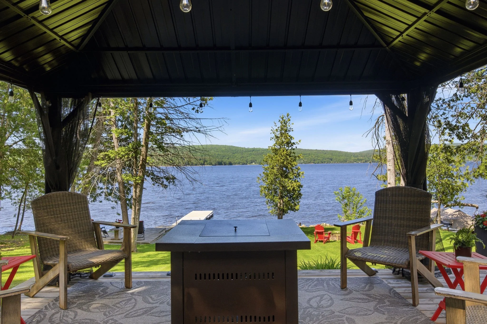 View from a backyard patio overlooking a lake with trees and hills in the distance. The patio has a large covered area with hanging string lights, outdoor chairs, and a fire pit table, creating a cozy outdoor seating area.
