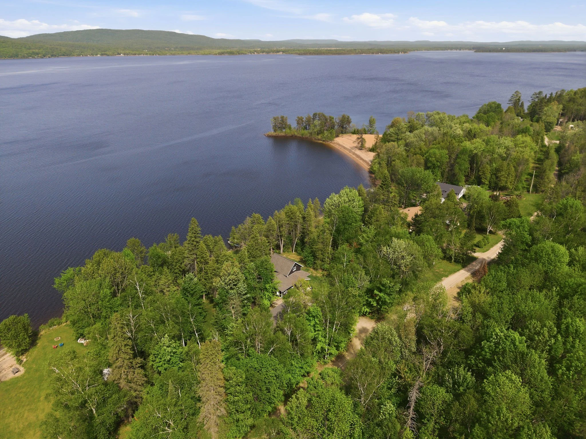 Aerial view of a large lake surrounded by lush green trees and some houses, with hills in the background under a partly cloudy sky.