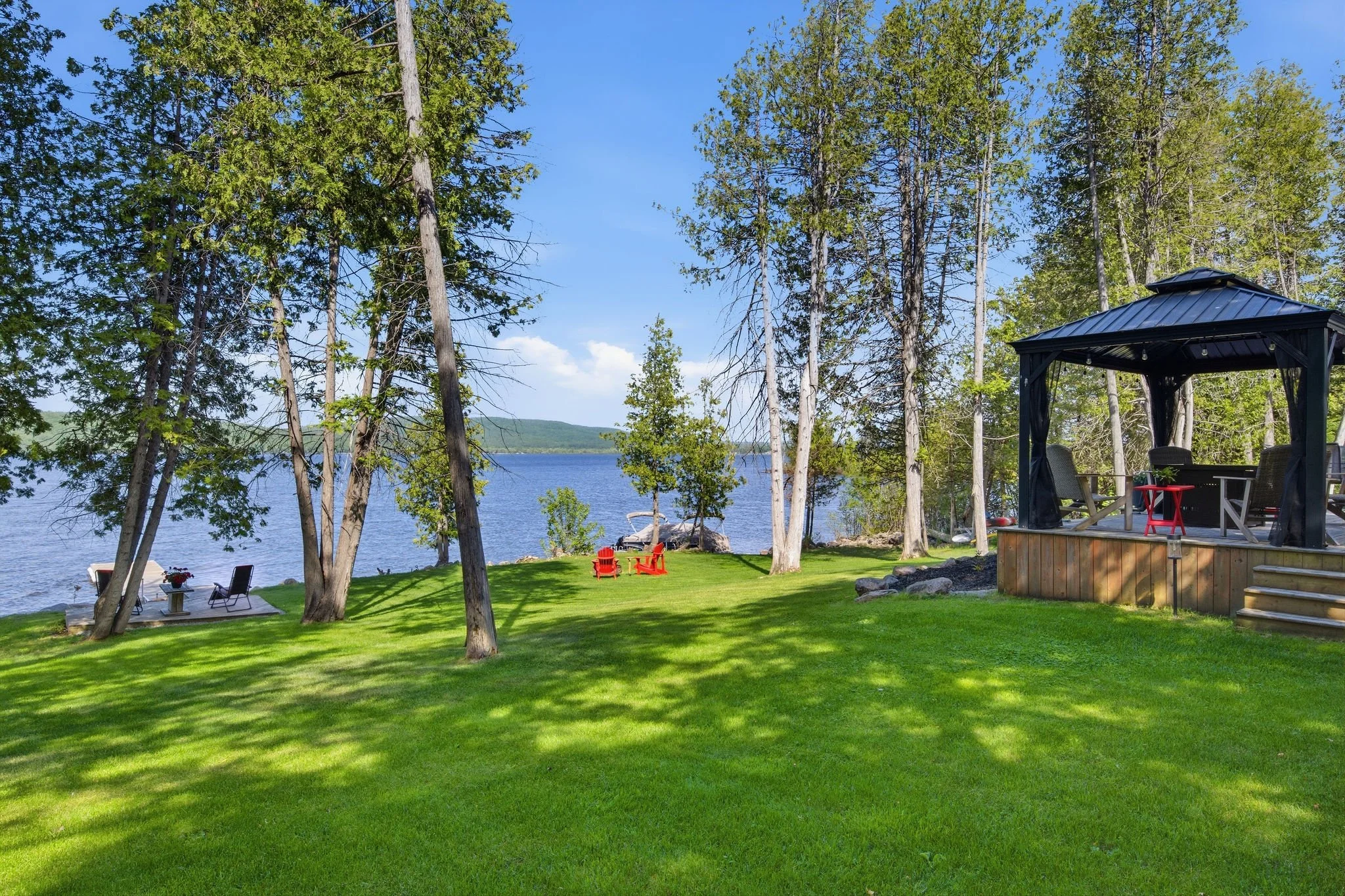 A lakeside backyard with lush green grass, surrounded by tall trees, overlooking a body of water with distant hills. There are outdoor chairs and a small table set up near the water, a covered gazebo with outdoor furniture, and a red Adirondack chair