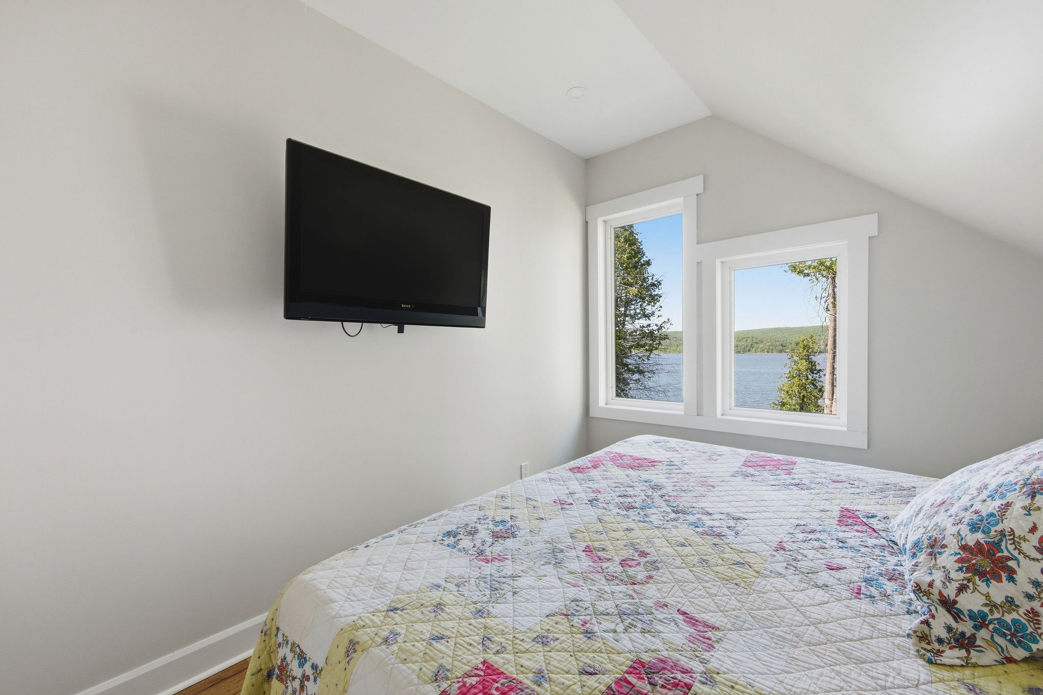 A bedroom with a bed covered in a quilt with a floral pattern, two windows showing trees and a lake outside, and a wall-mounted TV.