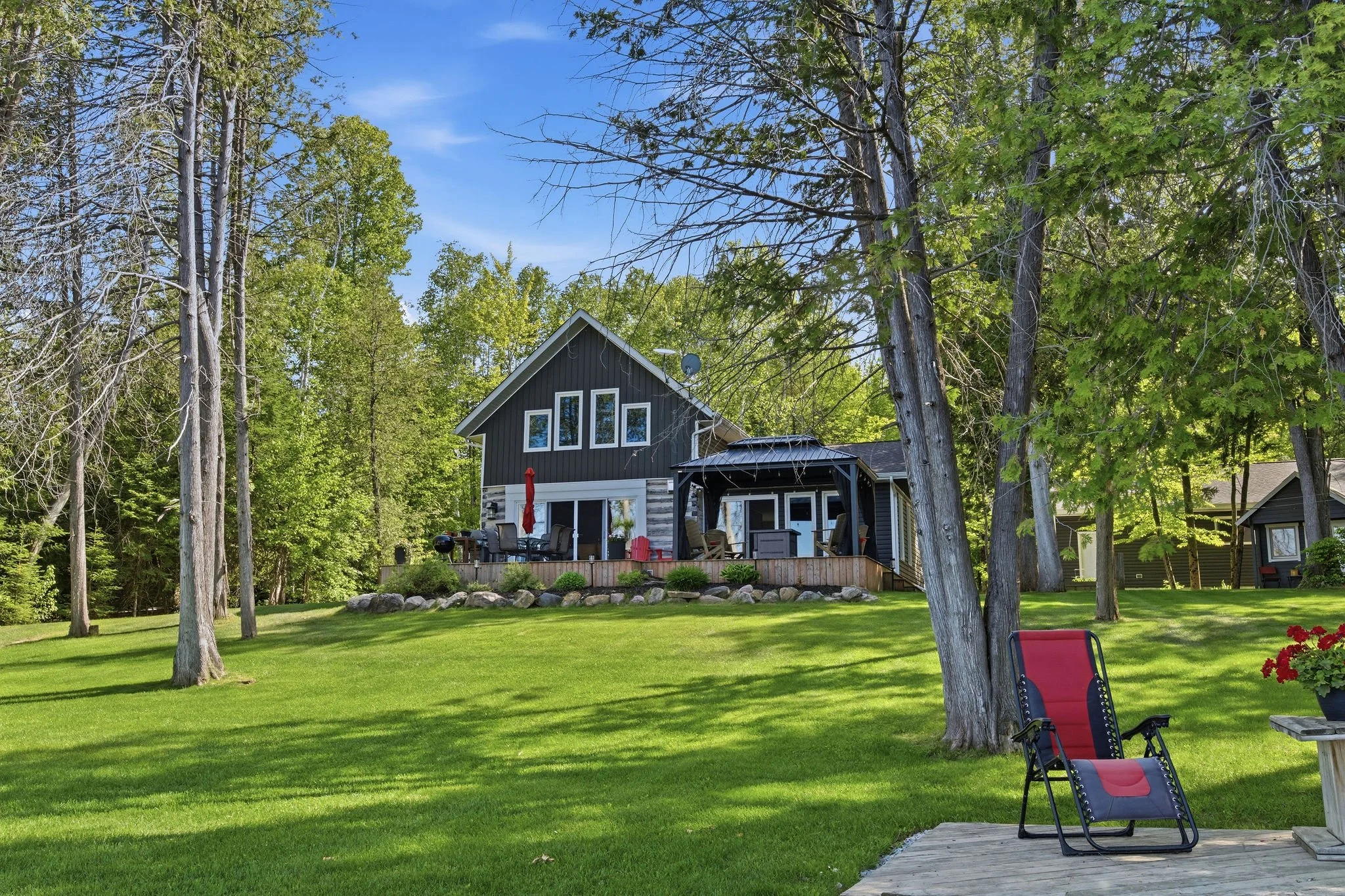 A backyard with green grass, tall trees, and a house with black and white exterior. There is a patio with outdoor furniture and a red chair, surrounded by a wooden deck. The scene is sunny with a clear blue sky.