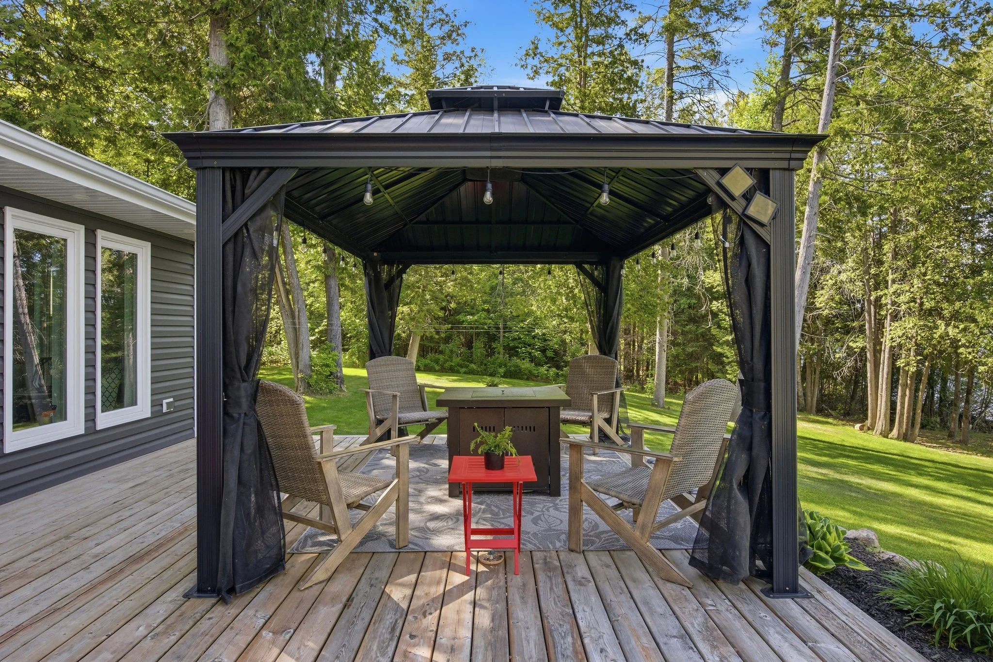 A backyard deck with a covered patio, surrounded by a green lawn and trees, furnished with chairs, a table, and hanging string lights.