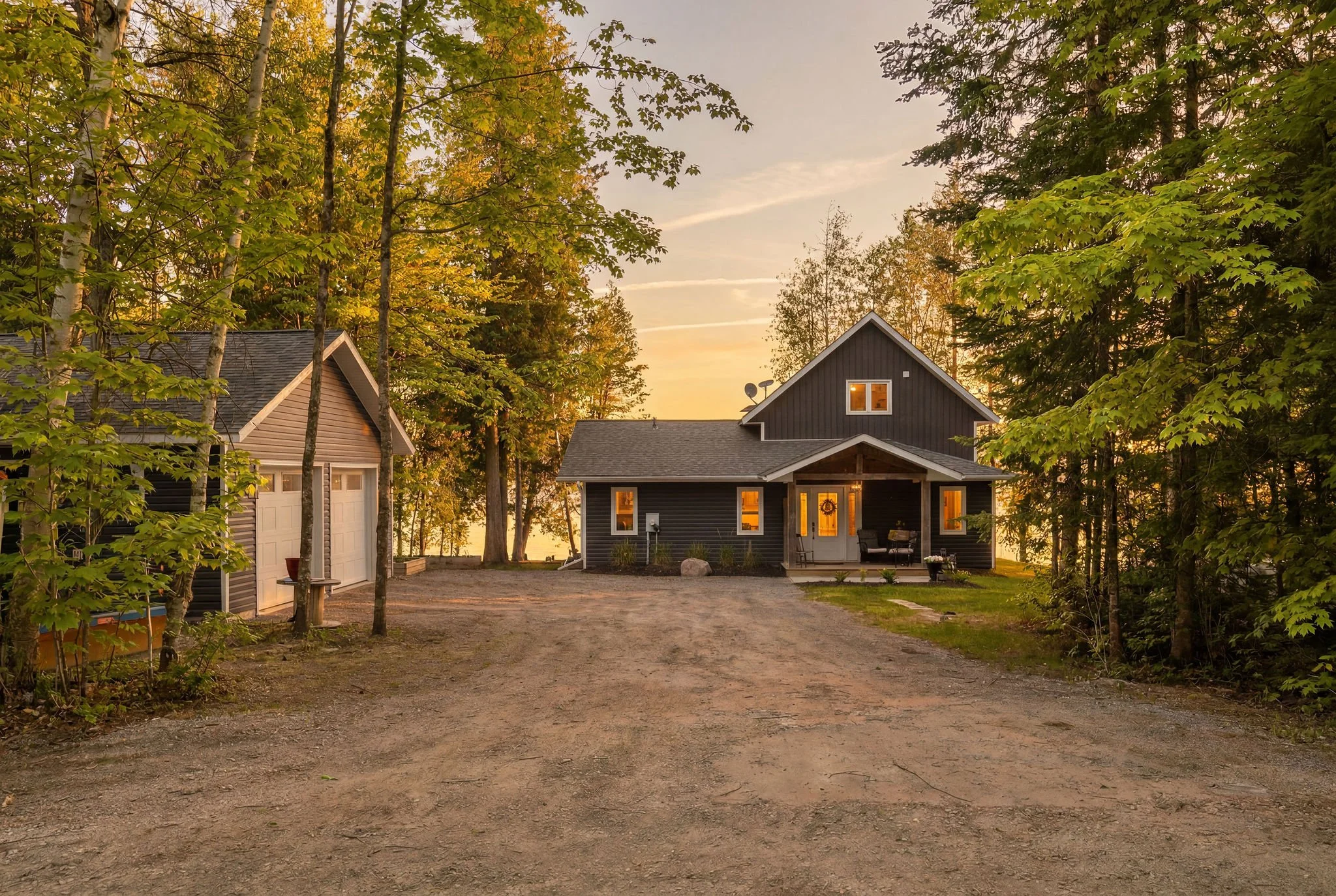 A house in a wooded area during sunset, with trees and a gravel driveway.