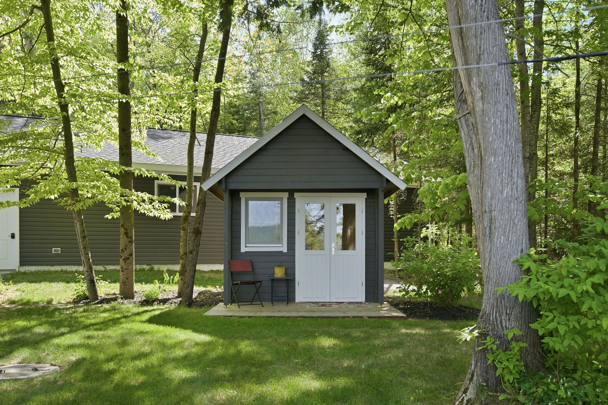 Small gray house with a gabled roof, white door, and a window, situated among trees with green leaves and a grassy lawn, in a wooded area.