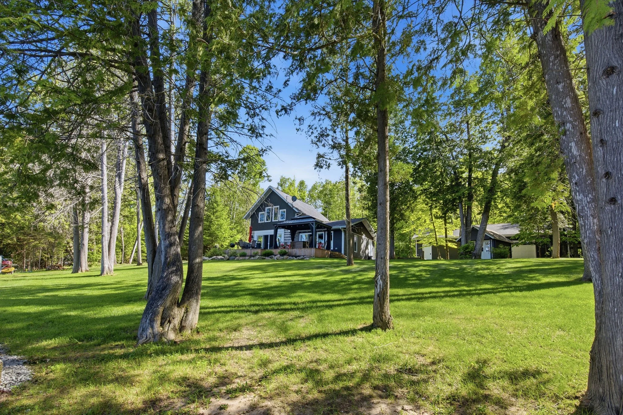 A backyard with lush green grass and tall trees surrounding a two-story house with dark gray siding, white window frames, and a deck with outdoor furniture.