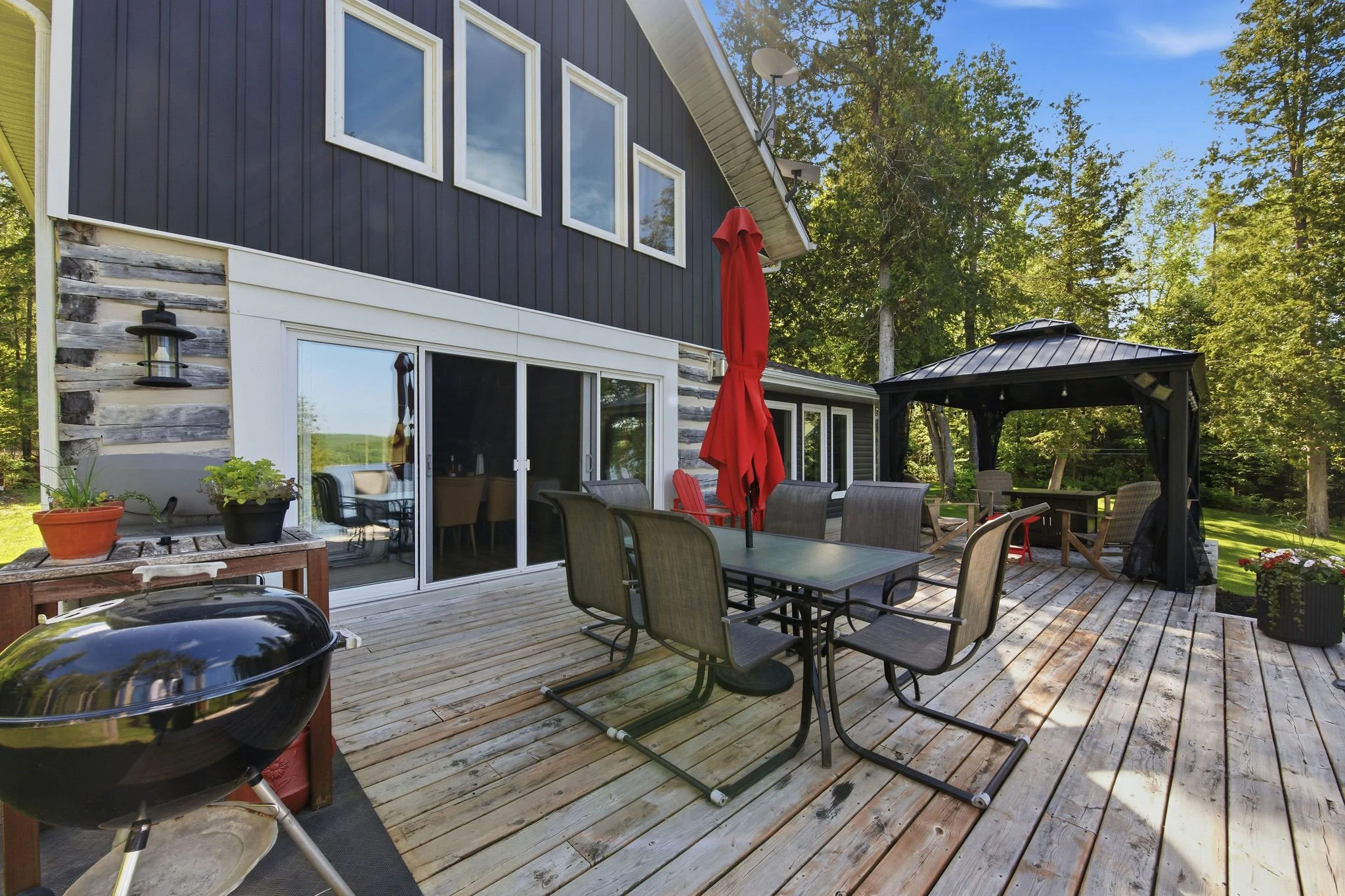 A wooden deck with outdoor furniture including a table, chairs, a red umbrella, and a grill. There is a black house with white trim, a sliding door, and nearby potted plants. A gazebo is in the background, surrounded by trees and greenery under a blu