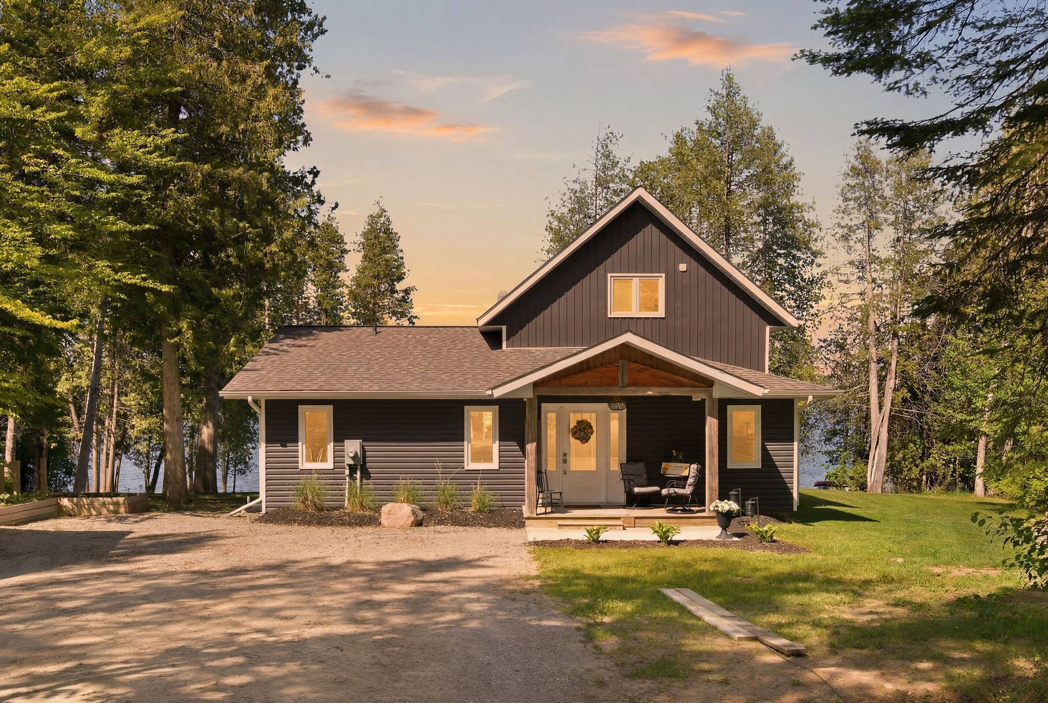 A two-story house with dark gray siding, white trim, and a front porch with seating, set among tall green trees with sunset clouds in the sky.