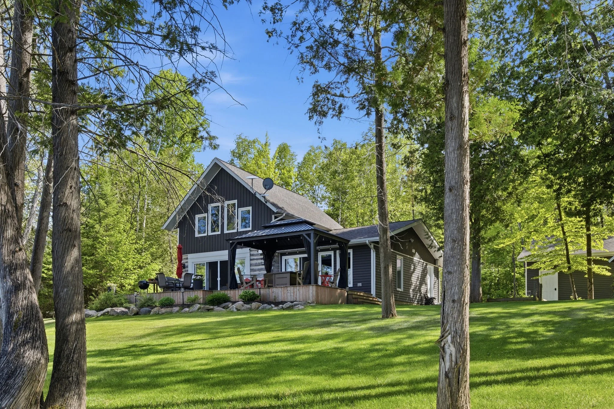 A modern house with large windows and an outdoor deck surrounded by a lush green yard and tall trees under a blue sky.