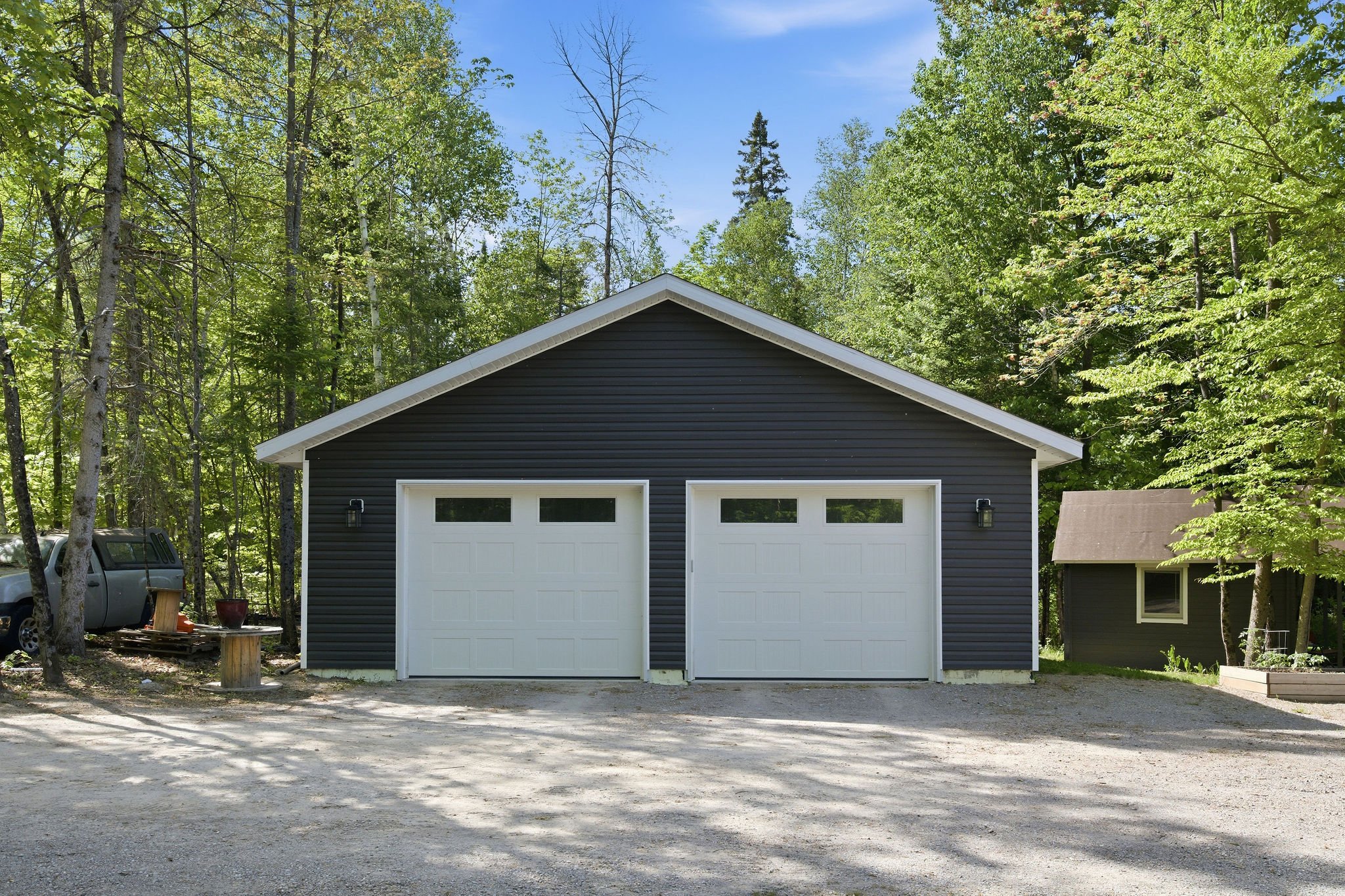 A black garage with white doors is situated in a wooded area with tall green trees surrounding it. A gravel driveway is in front of the garage, and a small shed is visible to the right.