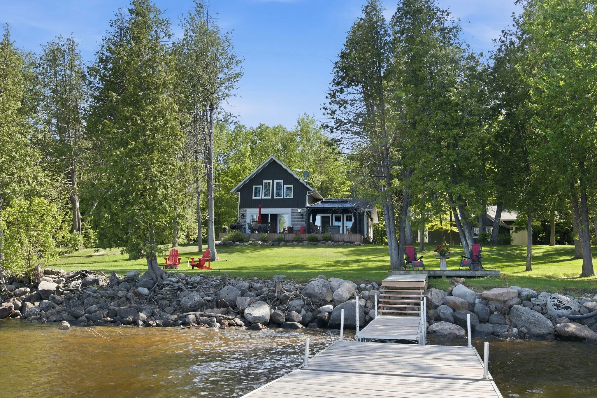 A lakeside backyard with a dock extending into the water, rocky shoreline, and a house surrounded by tall trees, with chairs and a table on the lawn.