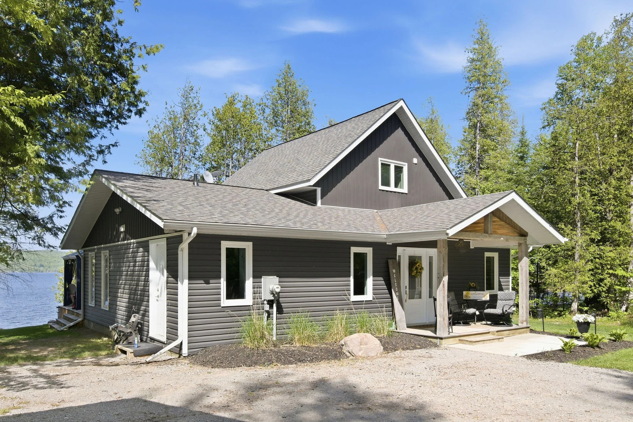 A modern gray house with white trim is surrounded by trees, with a gravel driveway in front and a lake visible to the left. There is outdoor furniture on the porch and decorative plants around the house.