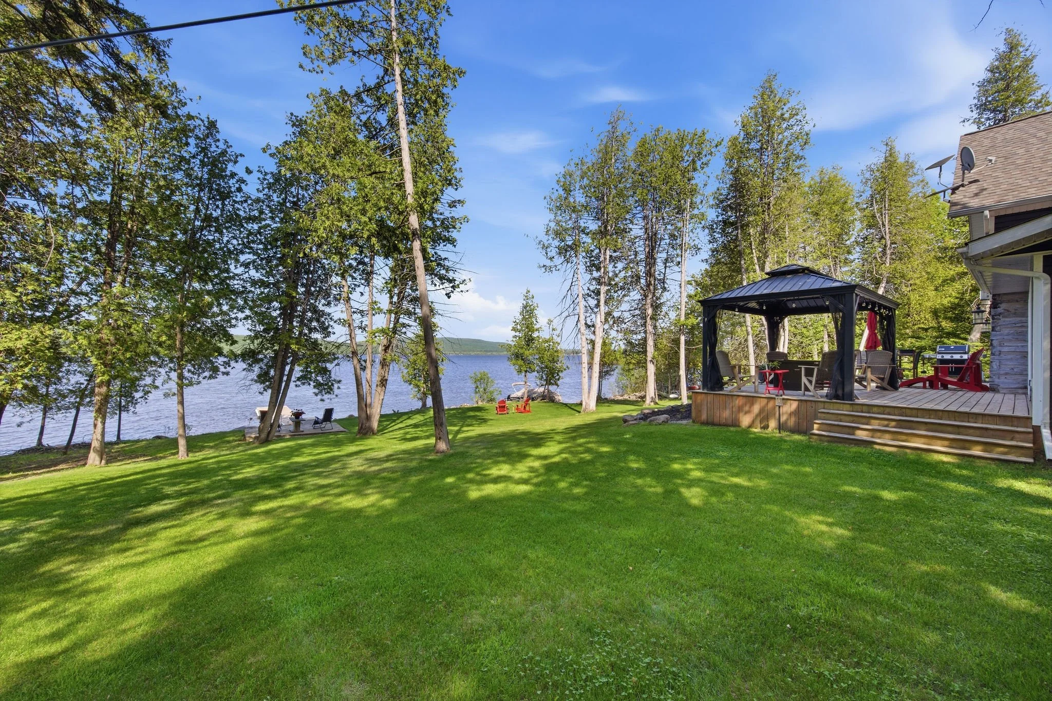 A backyard scene with a lush green lawn, tall trees, a lake in the background, and a house with a deck and outdoor furniture.