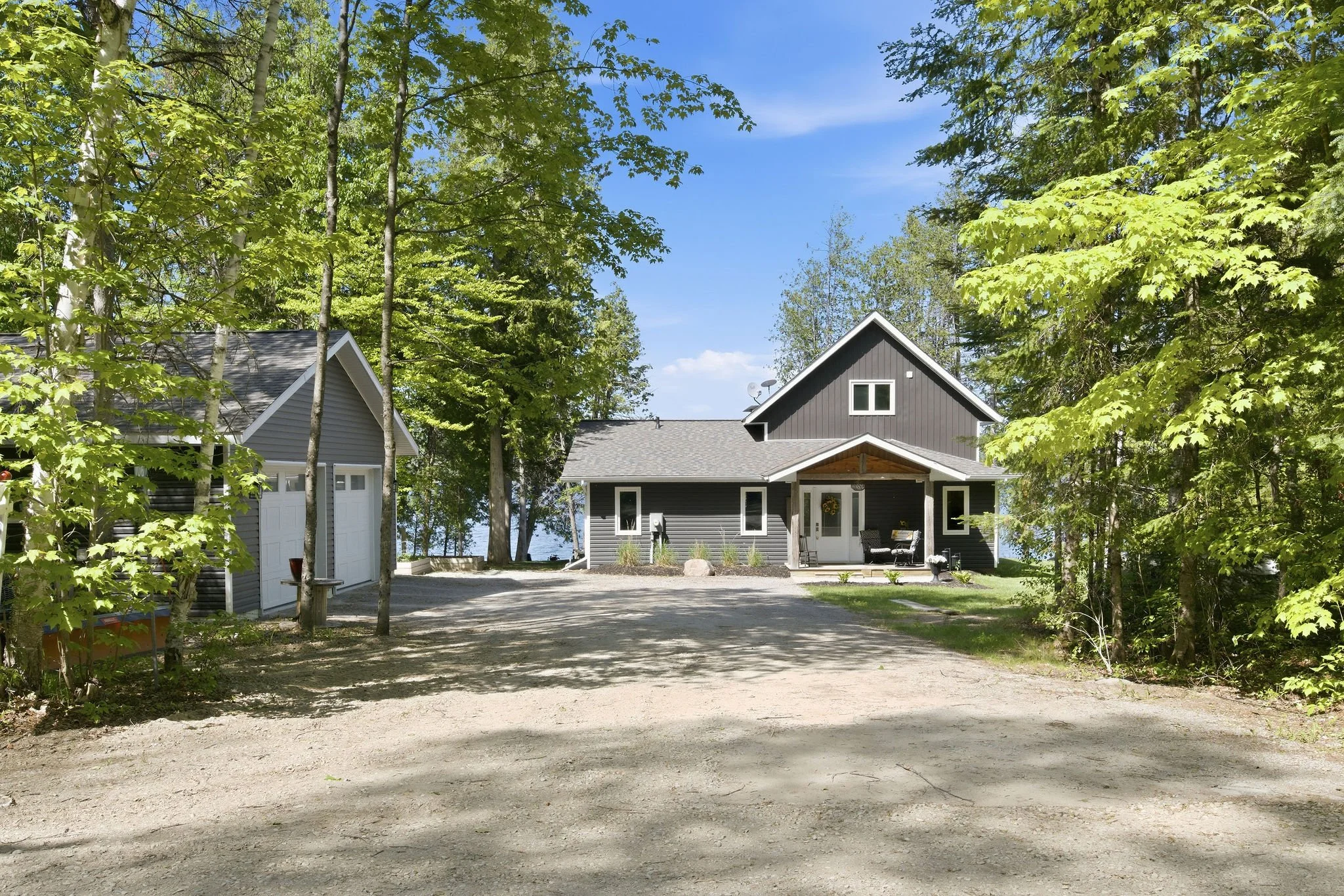 A house with a dark gray exterior and white trim, surrounded by tall green trees, with a gravel driveway in front and a view of a body of water in the background.