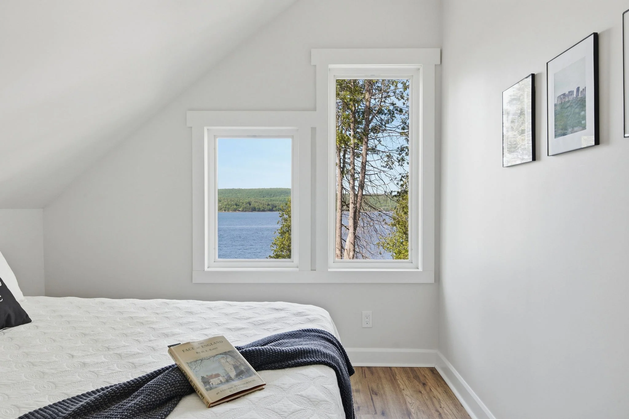 A bedroom with white walls, a bed with a white quilt, a book titled "Face of England" on the bed, and a large window showing a view of trees and water outside.