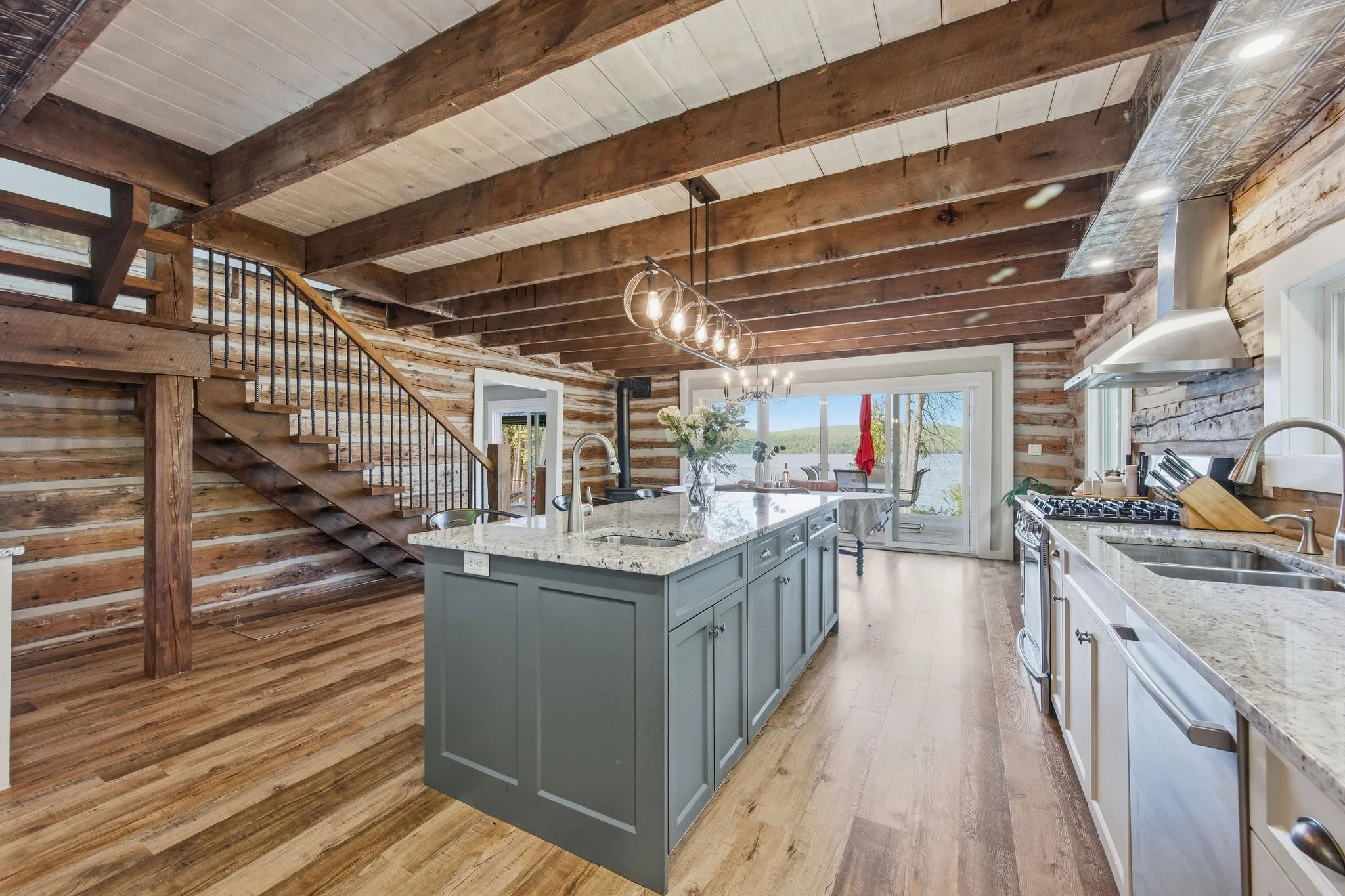 A rustic kitchen with wood-paneled walls and ceiling beams, an island with a granite countertop, pendant lighting, and large windows overlooking a lakeside view.