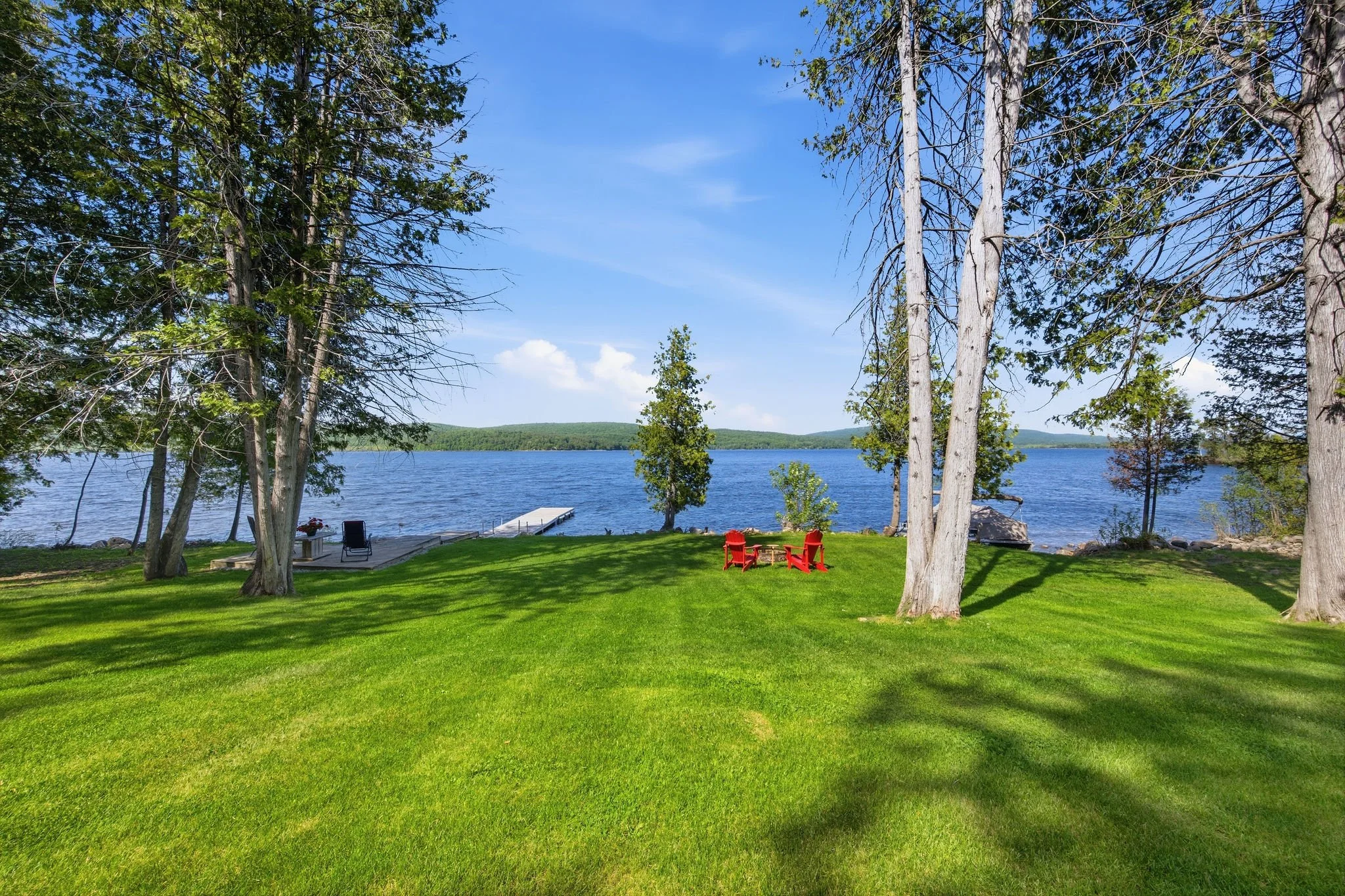 A lakeside yard with green grass, trees, and a dock leading to the water, featuring two red chairs and a small table.