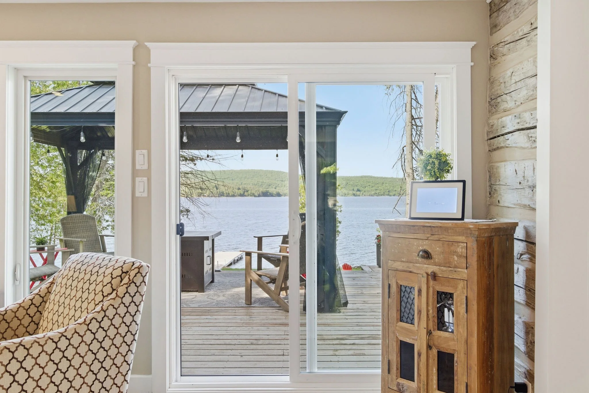 Room with a sliding glass door leading to a lakeside deck, featuring outdoor furniture and a view of the lake and green hills.