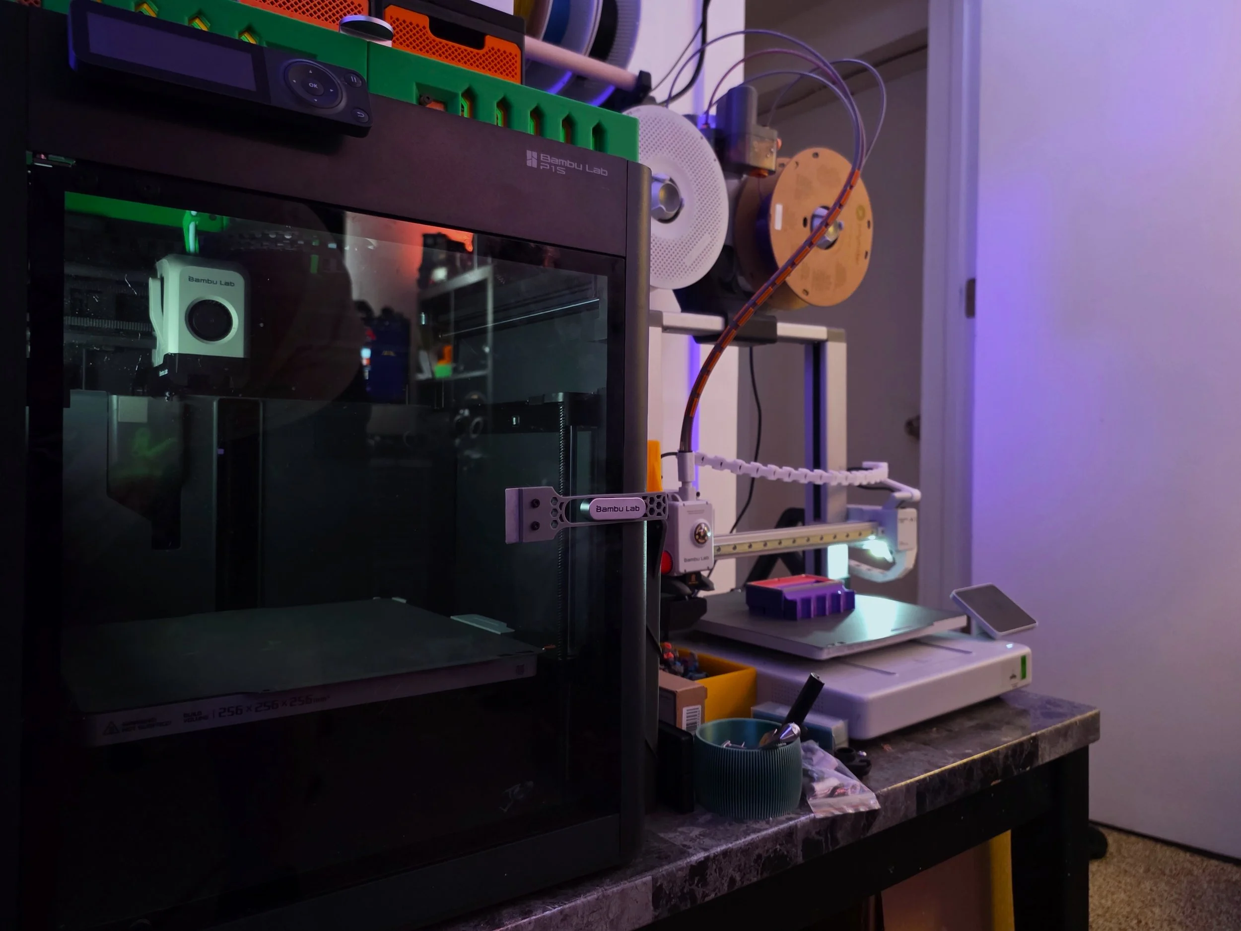 A cluttered workshop or lab space with electronic equipment, tools, and supplies on a countertop, including a Bambu Labs 3D printer, various wiring, and small containers for organizing parts.