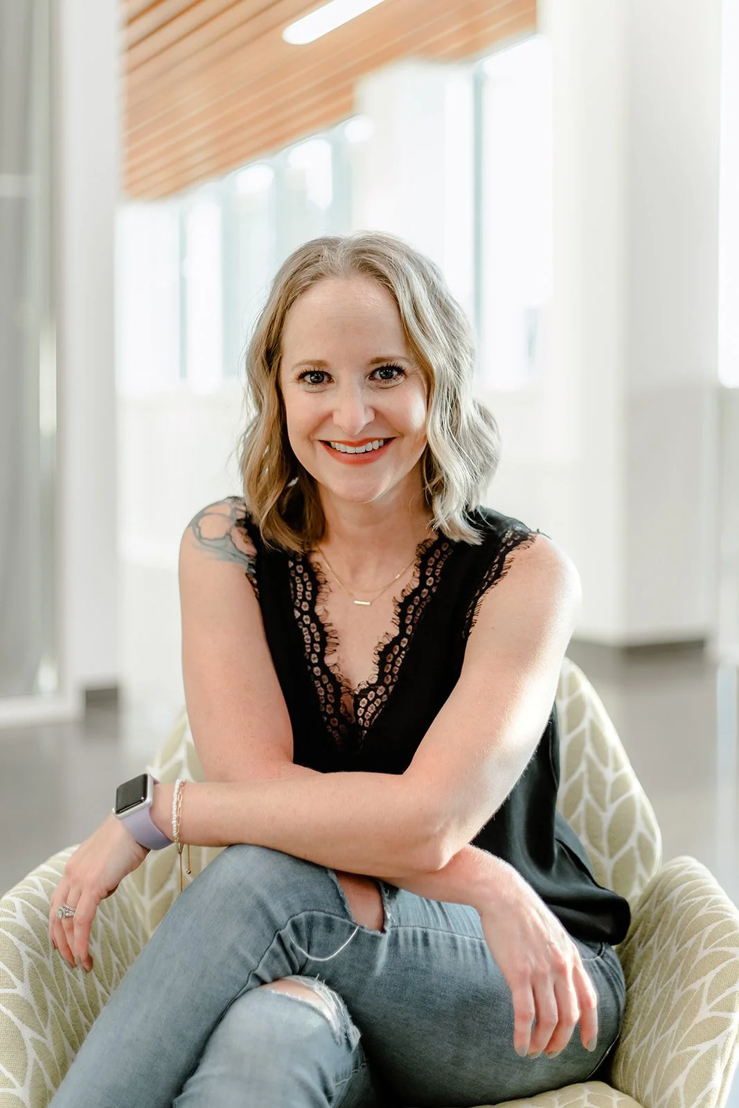 A smiling woman with blonde hair sitting on a patterned armchair in a bright room with large windows and a wooden ceiling.