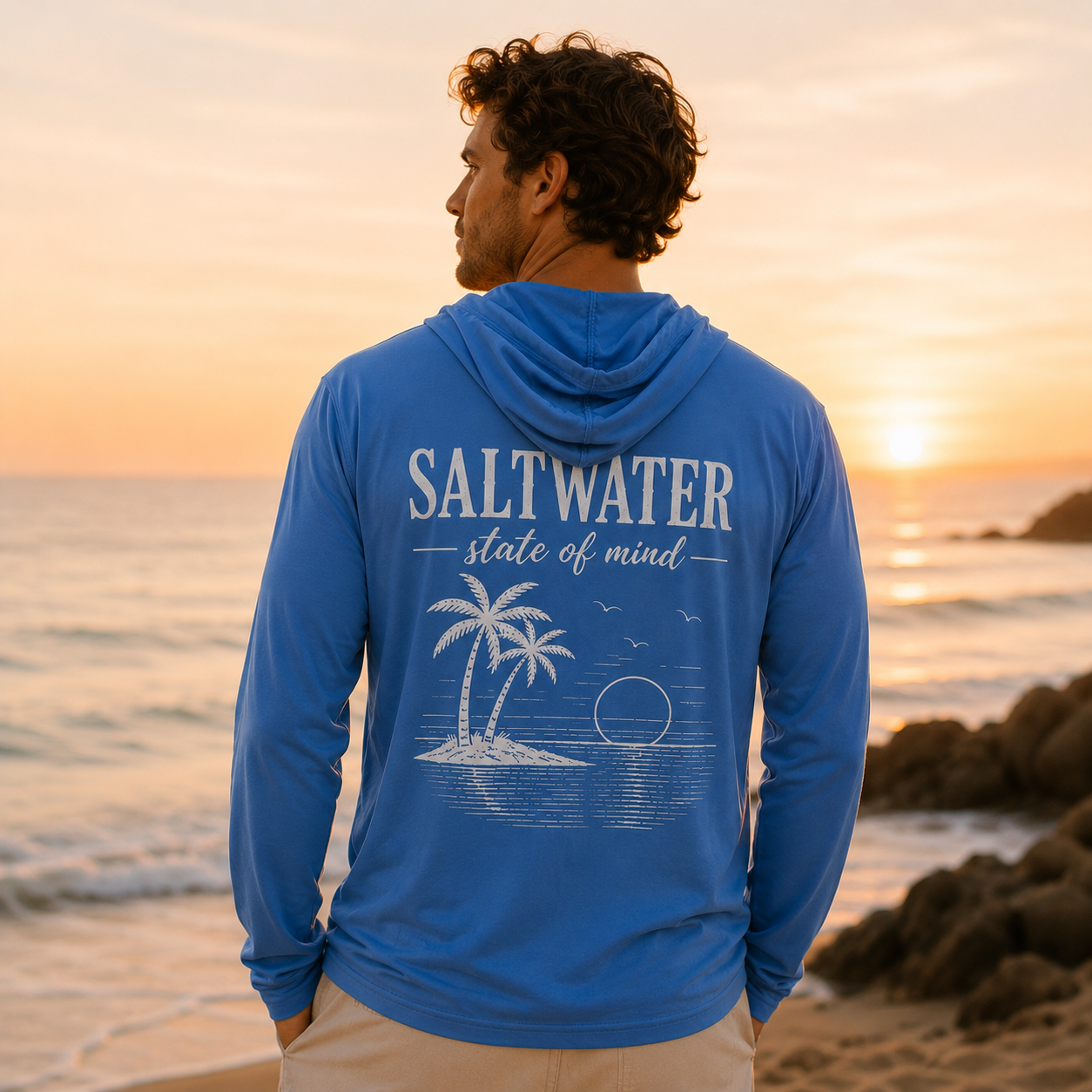 A man stands on a beach during sunset, wearing a blue hoodie with the words 'Saltwater, state of mind' and a graphic of palm trees and the ocean on the back.