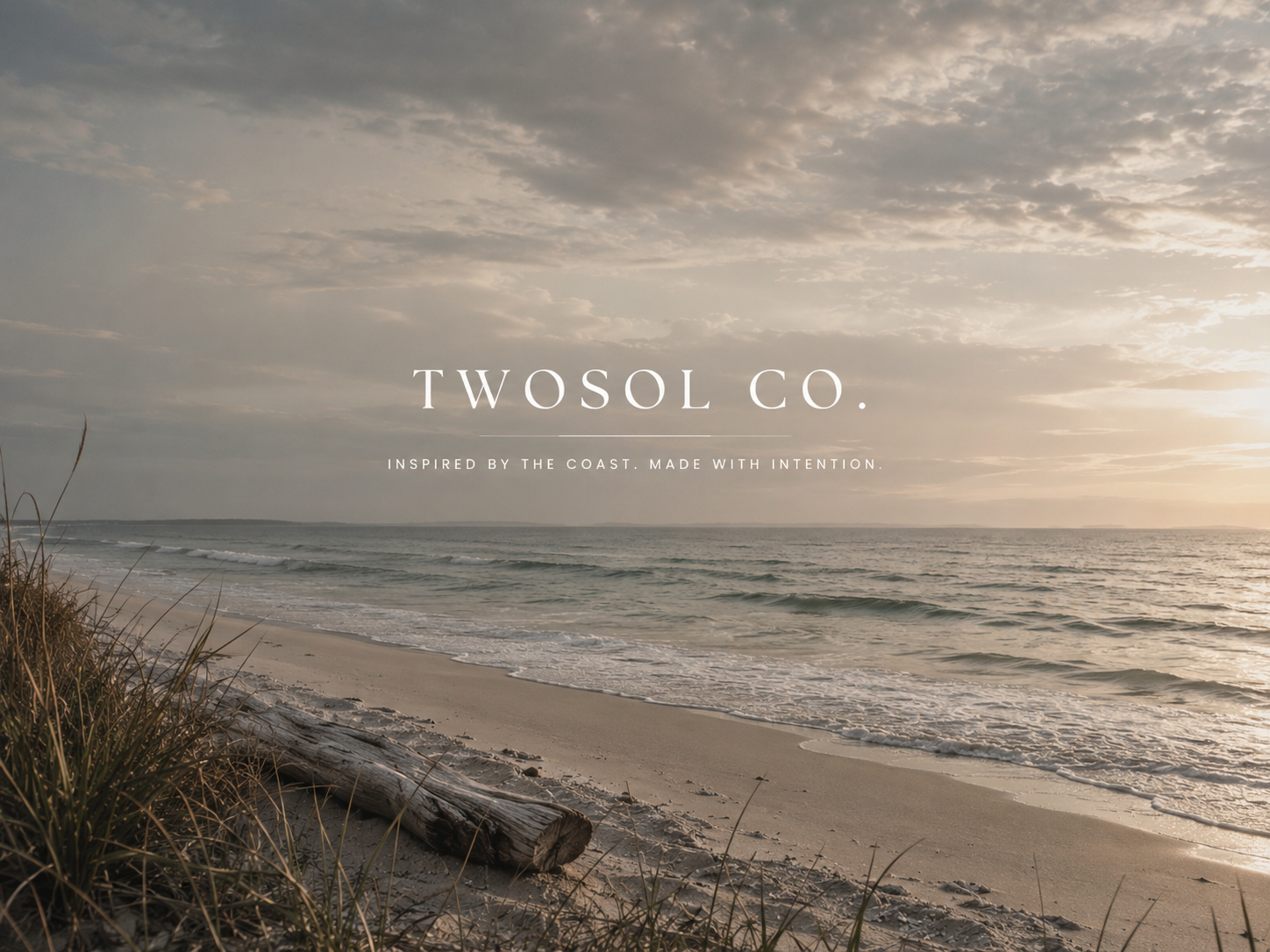 Sunset over a calm ocean with waves gently lapping against the sandy beach. There is some grass and driftwood in the foreground. The scene is serene and peaceful.