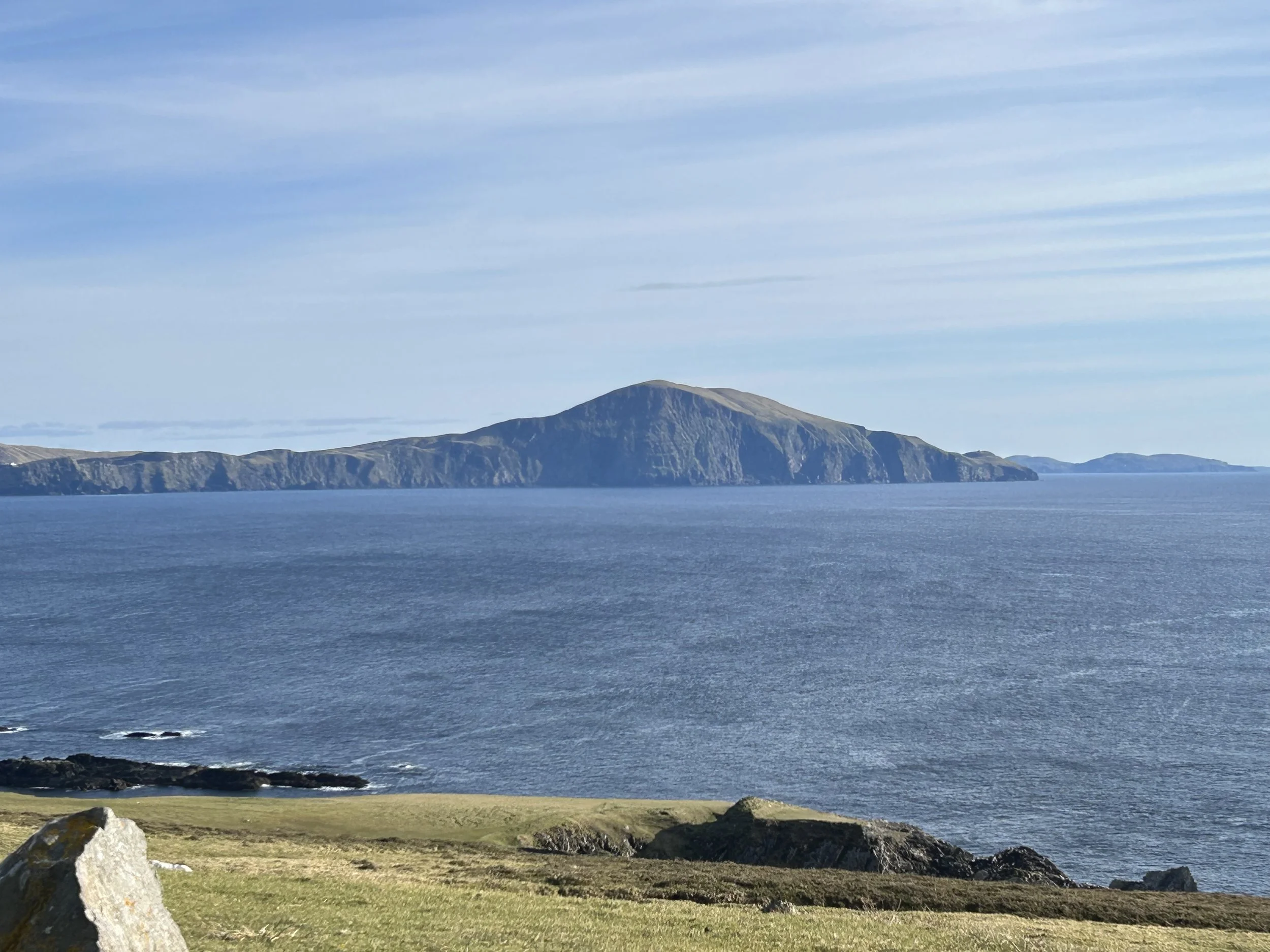 A scenic coastal landscape showing Clare Island in the background, surrounded by blue Atlantic waters and a grassy shoreline with rocks in the foreground.