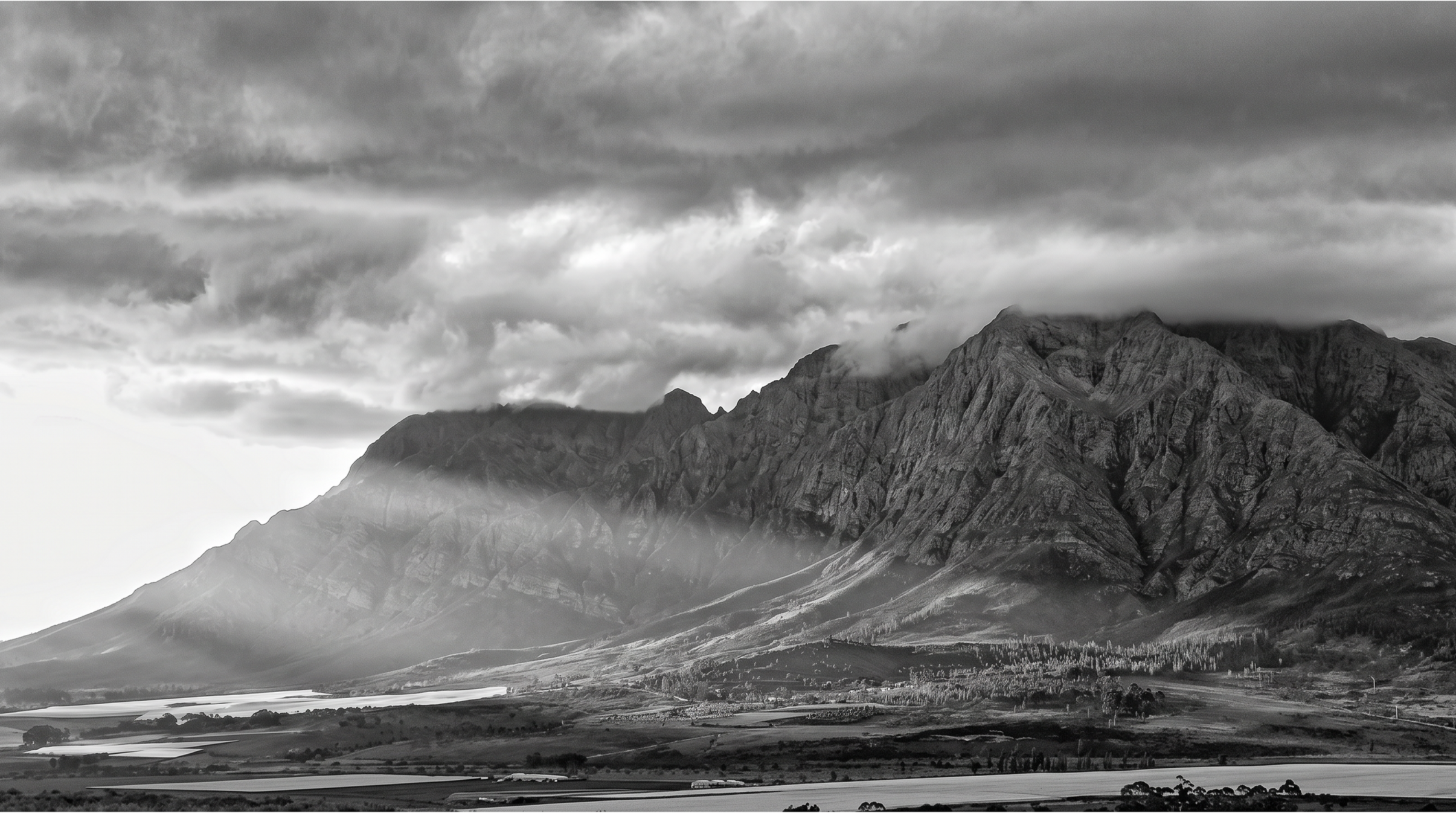 Black and white image of mountains with dramatic clouds above, a valley with fields, and a body of water in the foreground.
