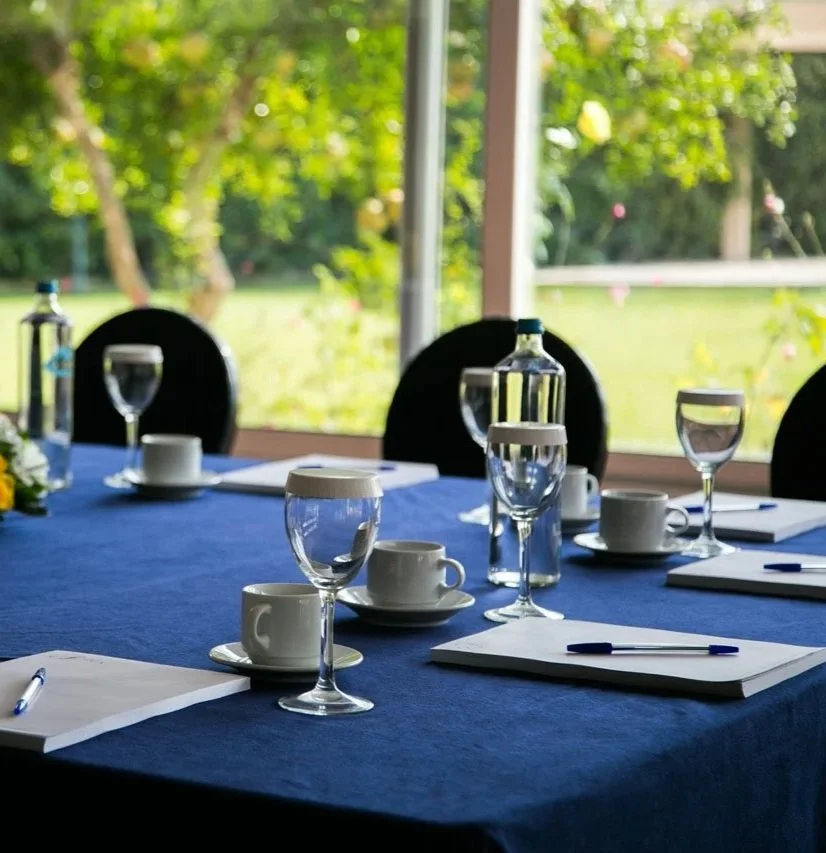 Meeting or conference table with notebooks, pens, water glasses, coffee cups, and bottles of water, set up for a discussion, with a view of greenery outside through large windows.