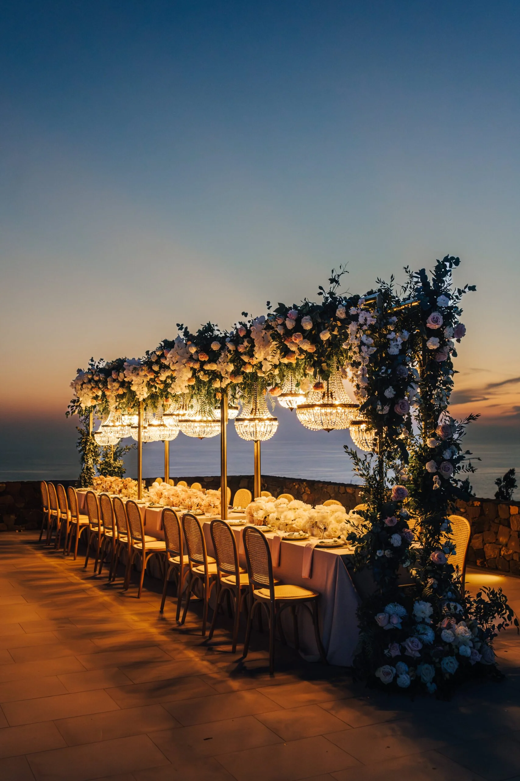 A long banquet table decorated with white and pink flowers and surrounded by chairs, set outdoors during sunset with a blue sky and calm ocean in the background. Overhead lighting and a floral arch add to the elegant setting, likely for a wedding or special event.