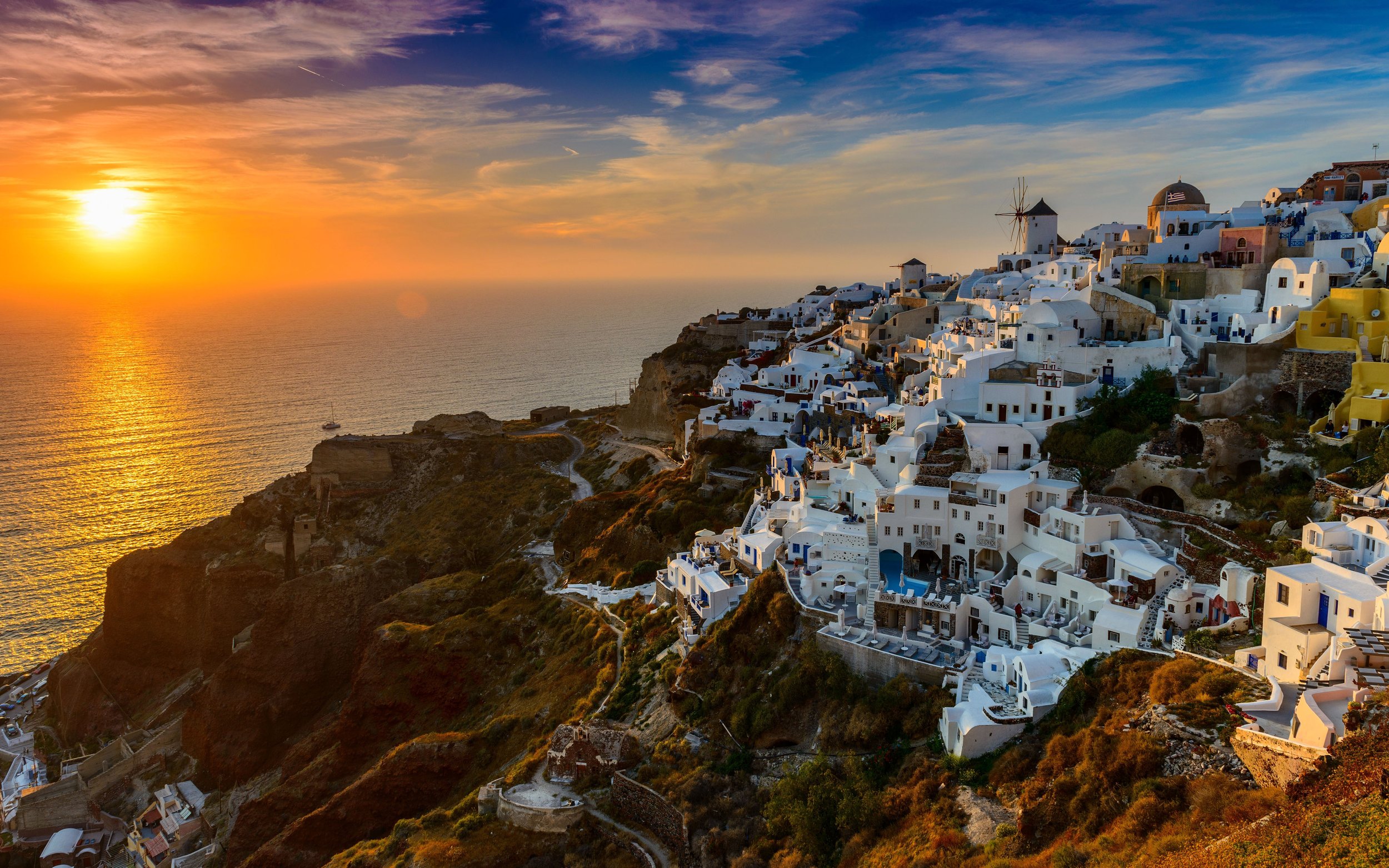 A coastal village on a hillside during sunset with white buildings, a windmill, and a calm sea.