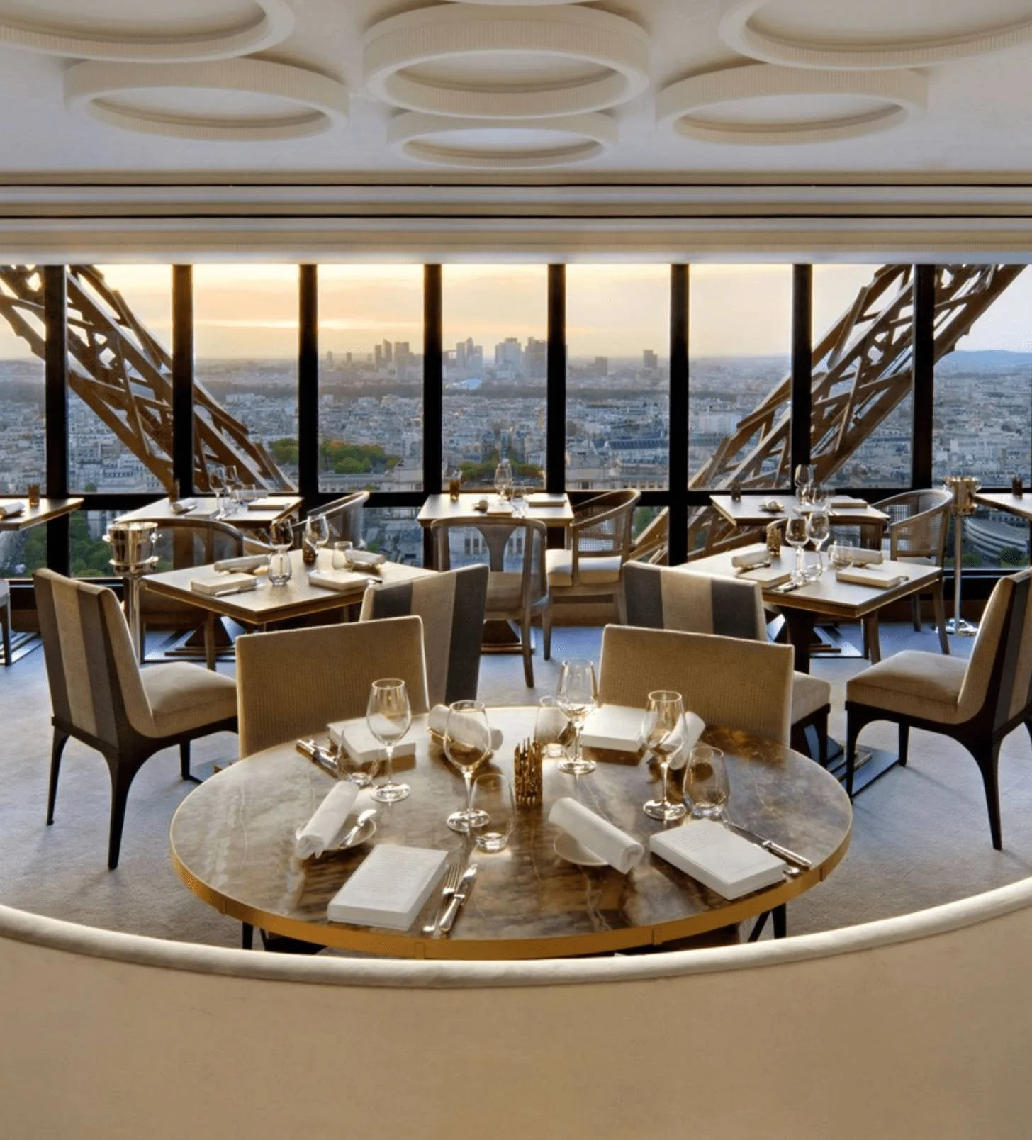 Elegant restaurant with set tables, wine glasses, and napkins, overlooking a city skyline through large windows.