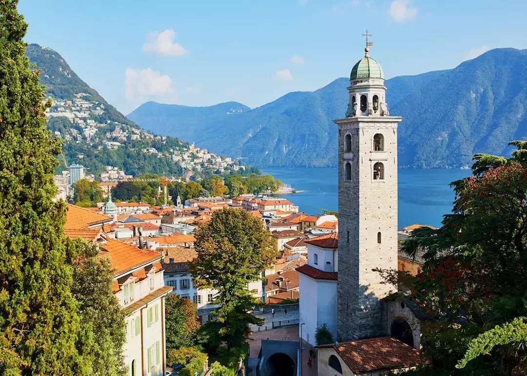 Tall historic clock tower with a green dome, overlooking a lakeside town with orange rooftops, surrounded by green trees and mountains in the background.