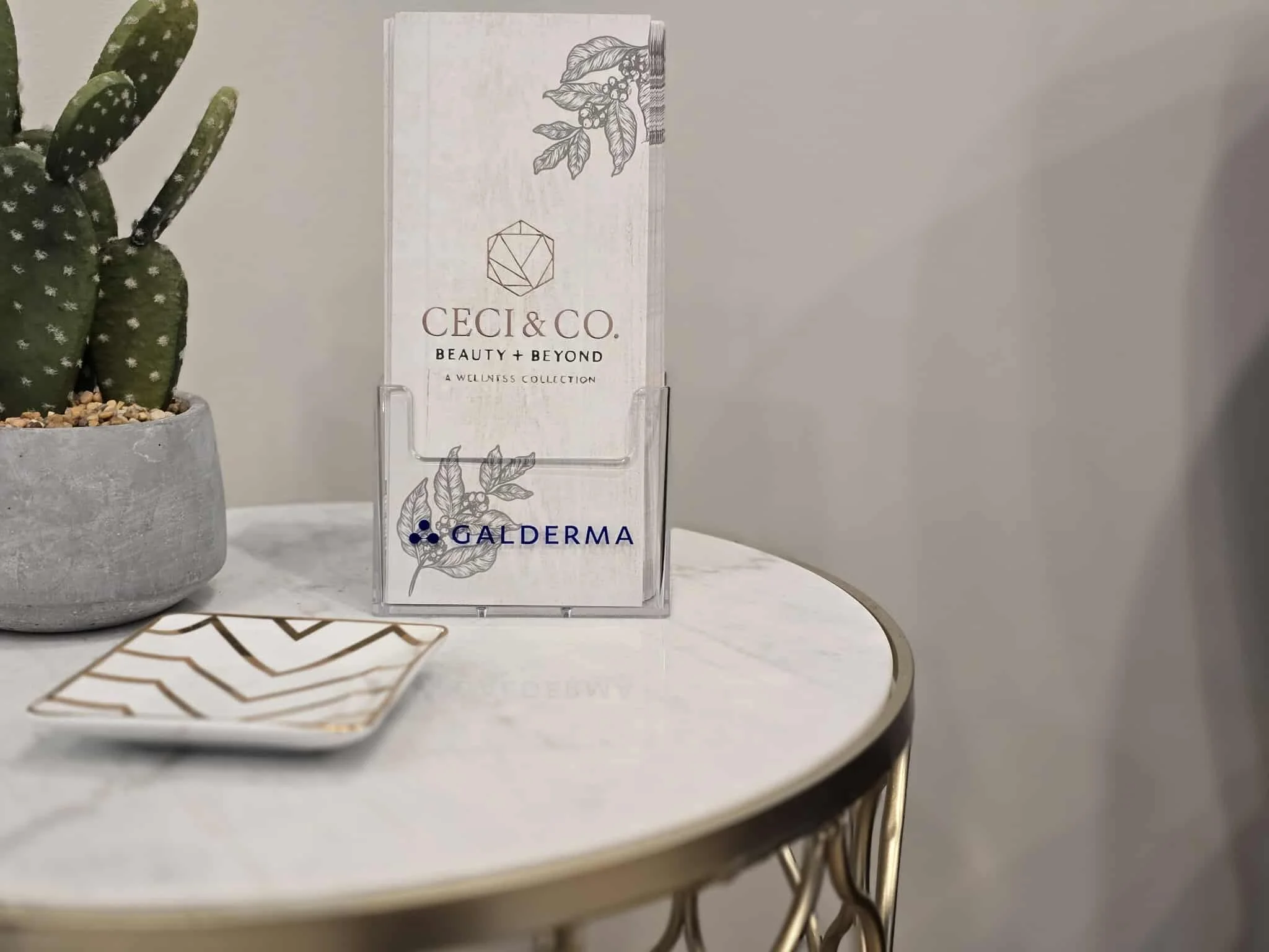 A white display card with botanical illustrations and the text "Ceci & Co. Beauty + Beyond A Wellness Collection" in brown, placed on a clear stand next to a potted cactus on a round white table with a gold edge.