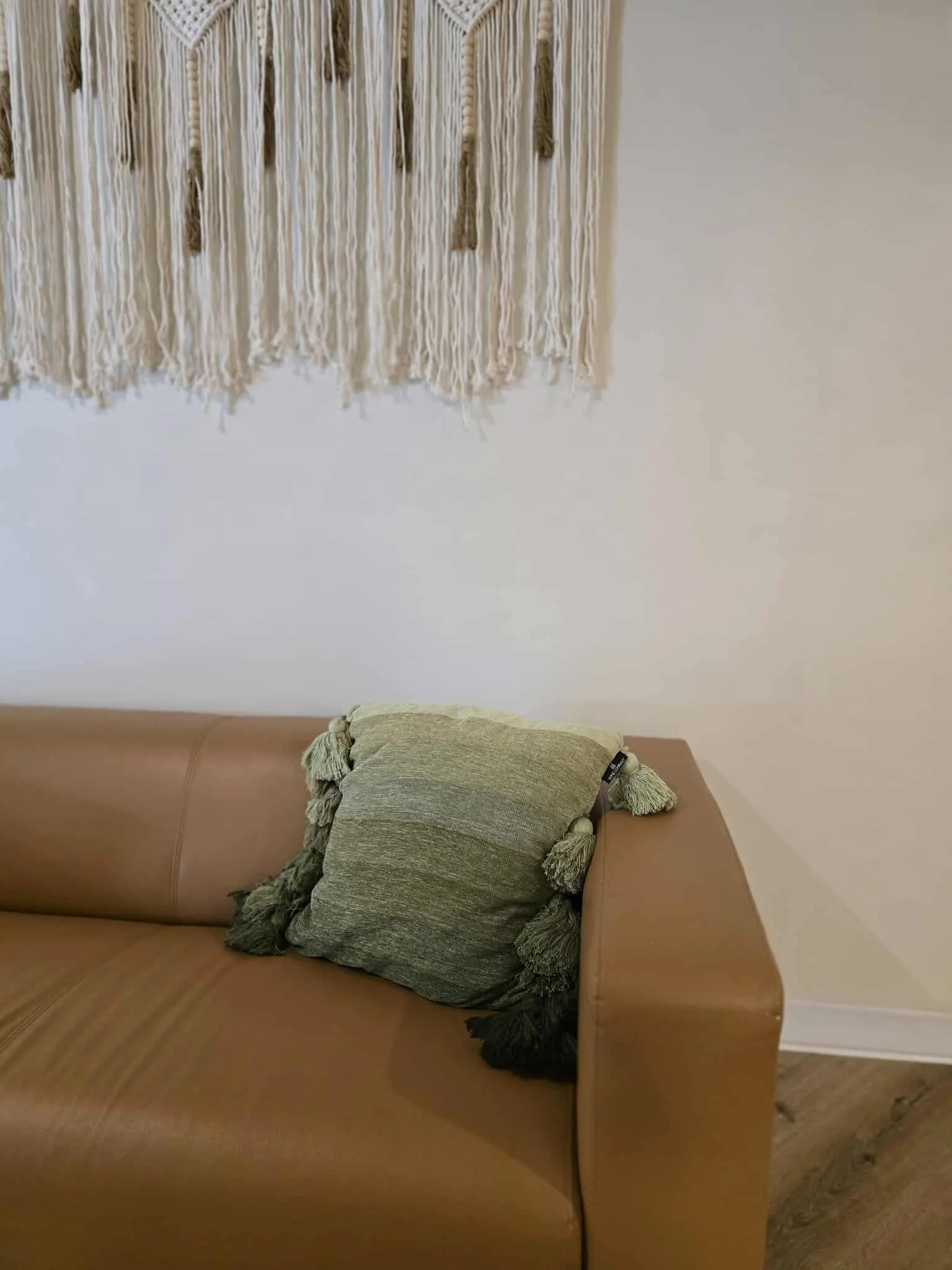 A tan sofa with a green pillow that has tassels on the corners, positioned in front of a white wall with a cream-colored woven wall hanging.