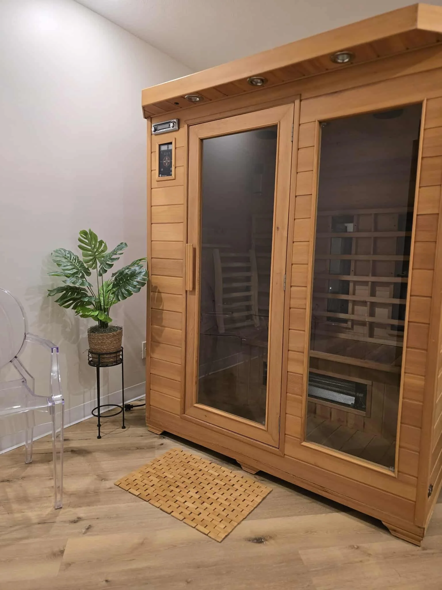 A wooden infrared sauna with a glass door in a room with light-colored walls and flooring. To the left, there is a potted plant on a small black stand, a clear acrylic chair, and a small mat on the floor.