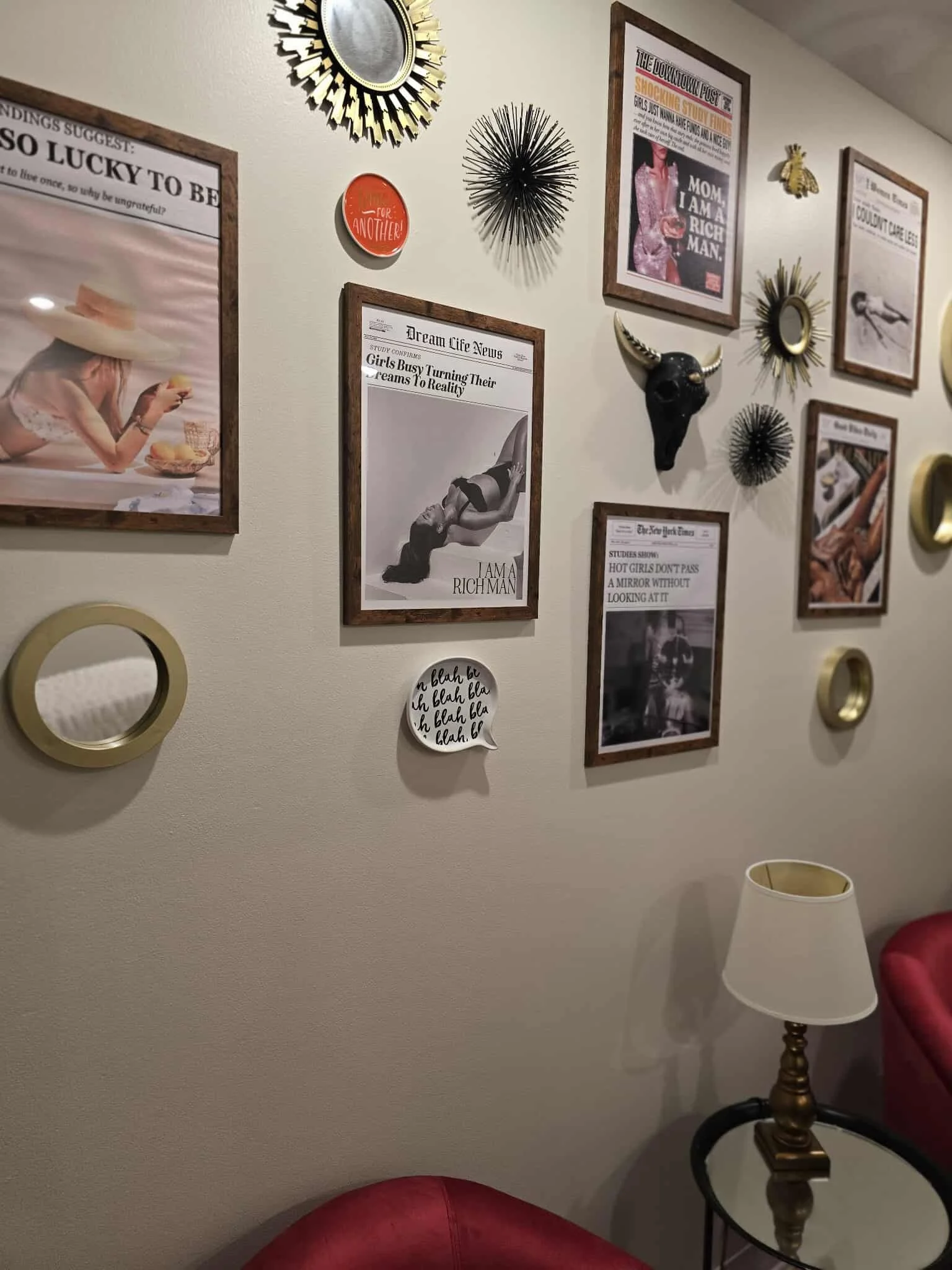 Decorative living room wall with framed magazine covers and art prints, round mirrors, and metallic wall sculptures. A side table with a traditional lamp and a red chair are partially visible.