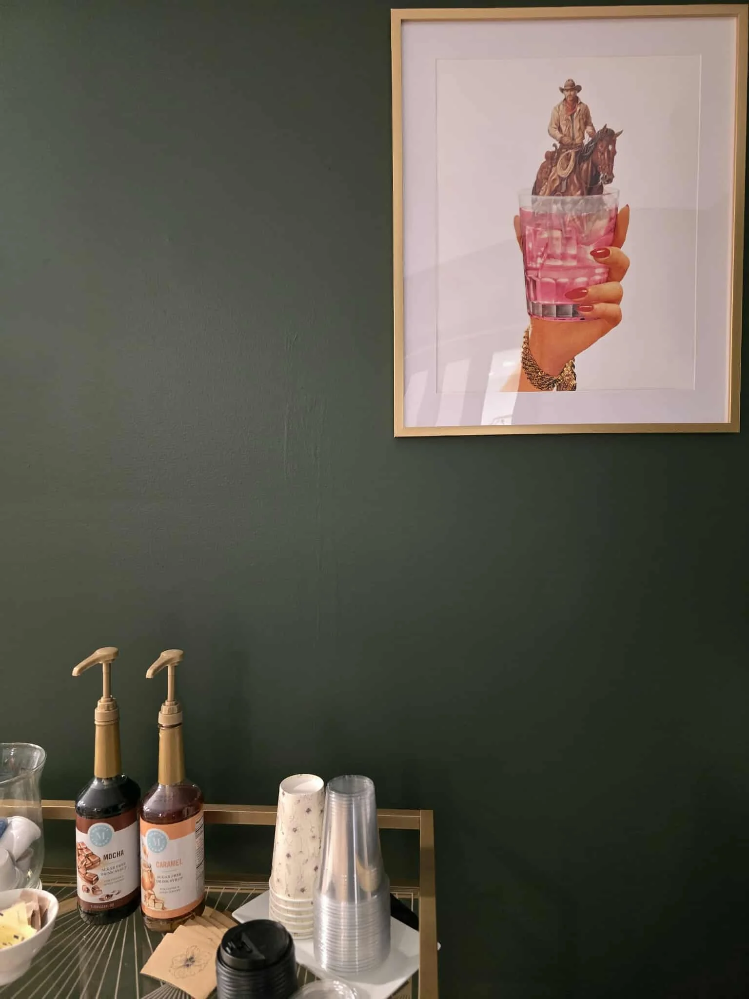 Framed artwork on dark green wall depicting a hand holding a glass of pink drink with a miniature cowboy riding a horse emerging from the glass.