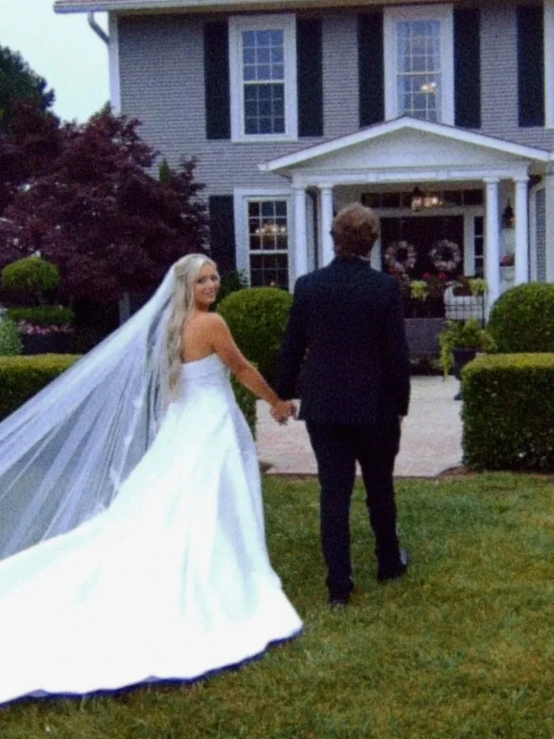 A bride with long blonde hair in a white wedding gown and veil holding hands with a groom in a suit outside a house. Taken on a disposable camera to give a nostalgic, vintage look.