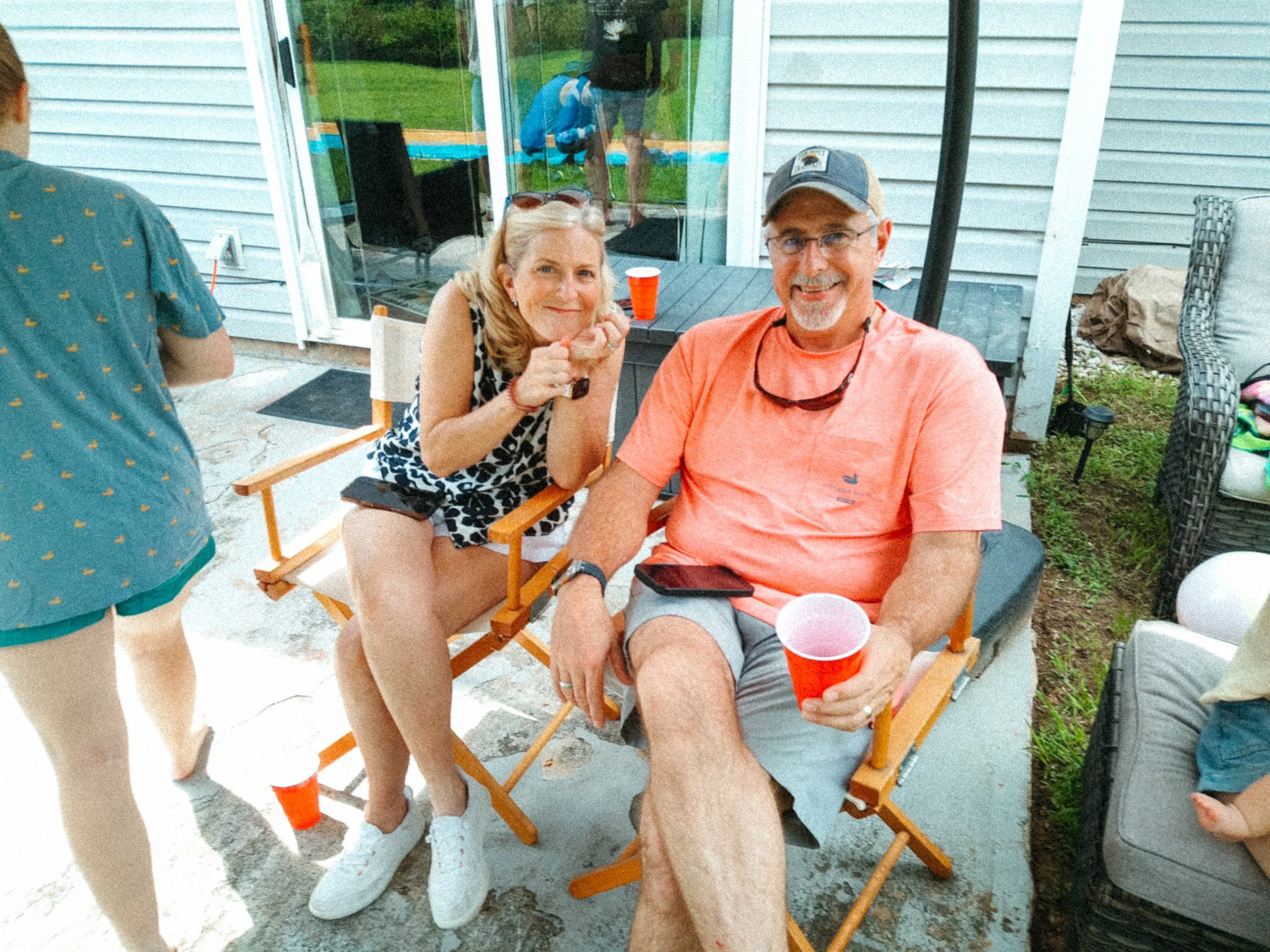 A smiling man in a gray cap and orange shirt holding a red cup, sitting on a wooden chair outdoors. A woman with blonde hair and sunglasses on her head is leaning in toward him, smiling. They are sitting at a table on a porch with a sliding glass door and a person visible inside. There is a person in a teal shirt with yellow patterns standing on the left. Backyard and greenery are visible through the glass door.
