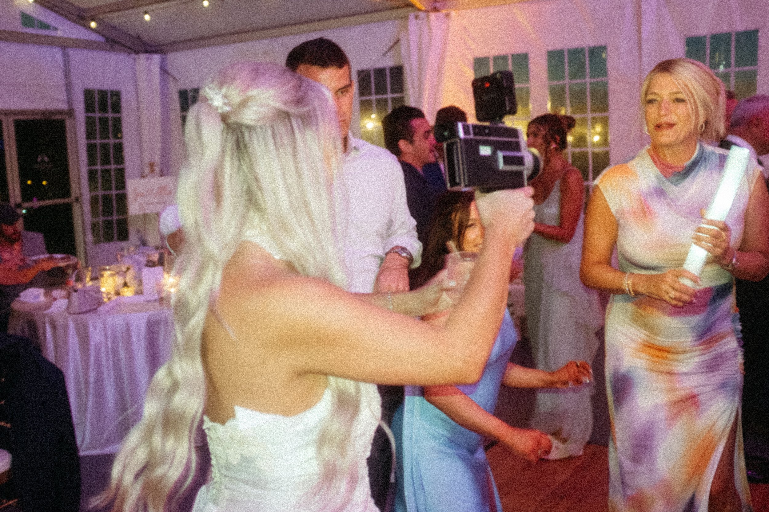 A group of people at a social event in a tented area with string lights, including a woman wearing a colorful dress holding a glow stick, a man with short dark hair and a white shirt, and a woman with long blonde hair with floral accessories, taking pictures of each other.