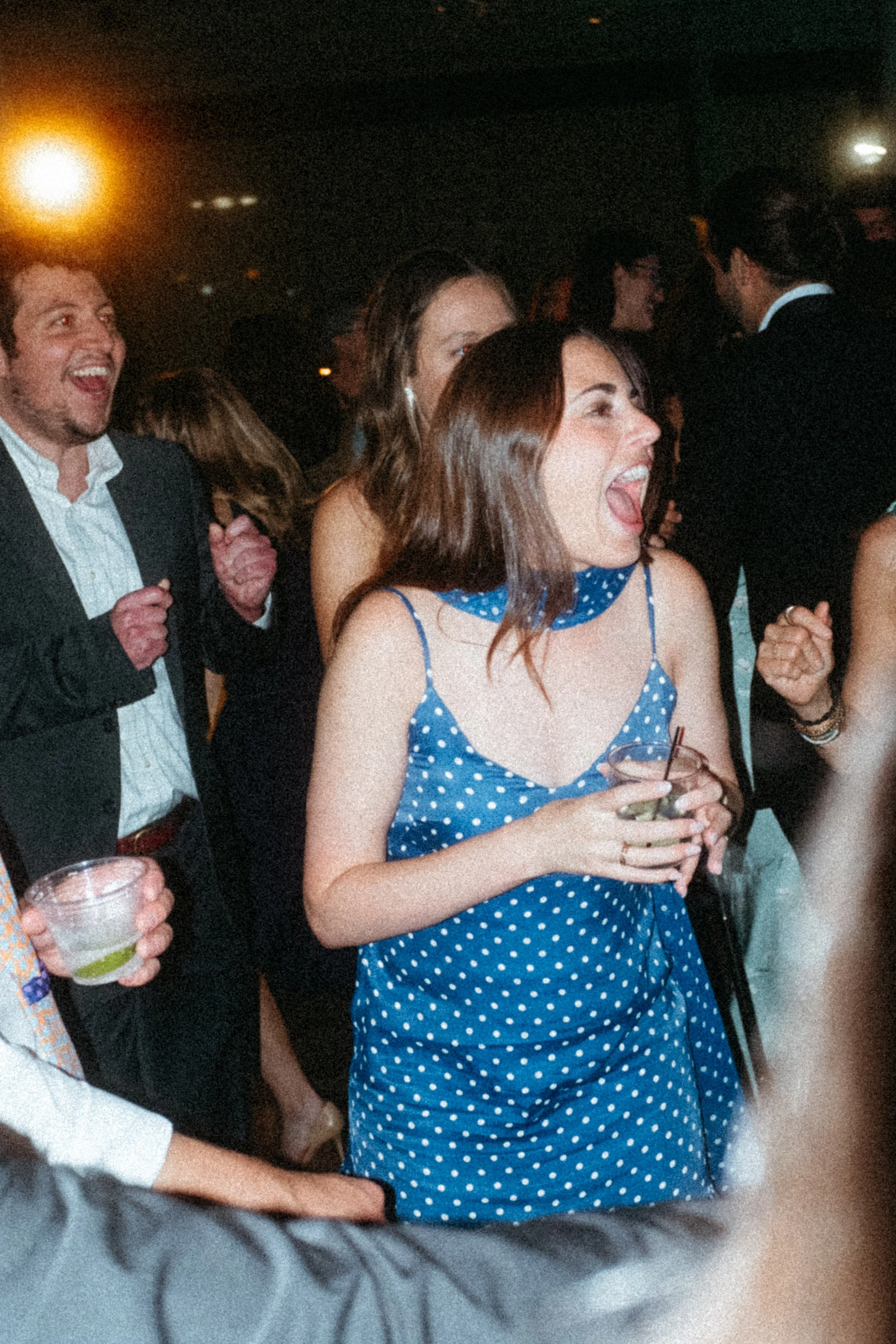A woman in a blue polka dot dress laughing and holding a drink at a party. Others around her are also enjoying themselves, with some smiling and dancing. The background shows a dark indoor setting with warm lighting. Taken on a disposable camera to