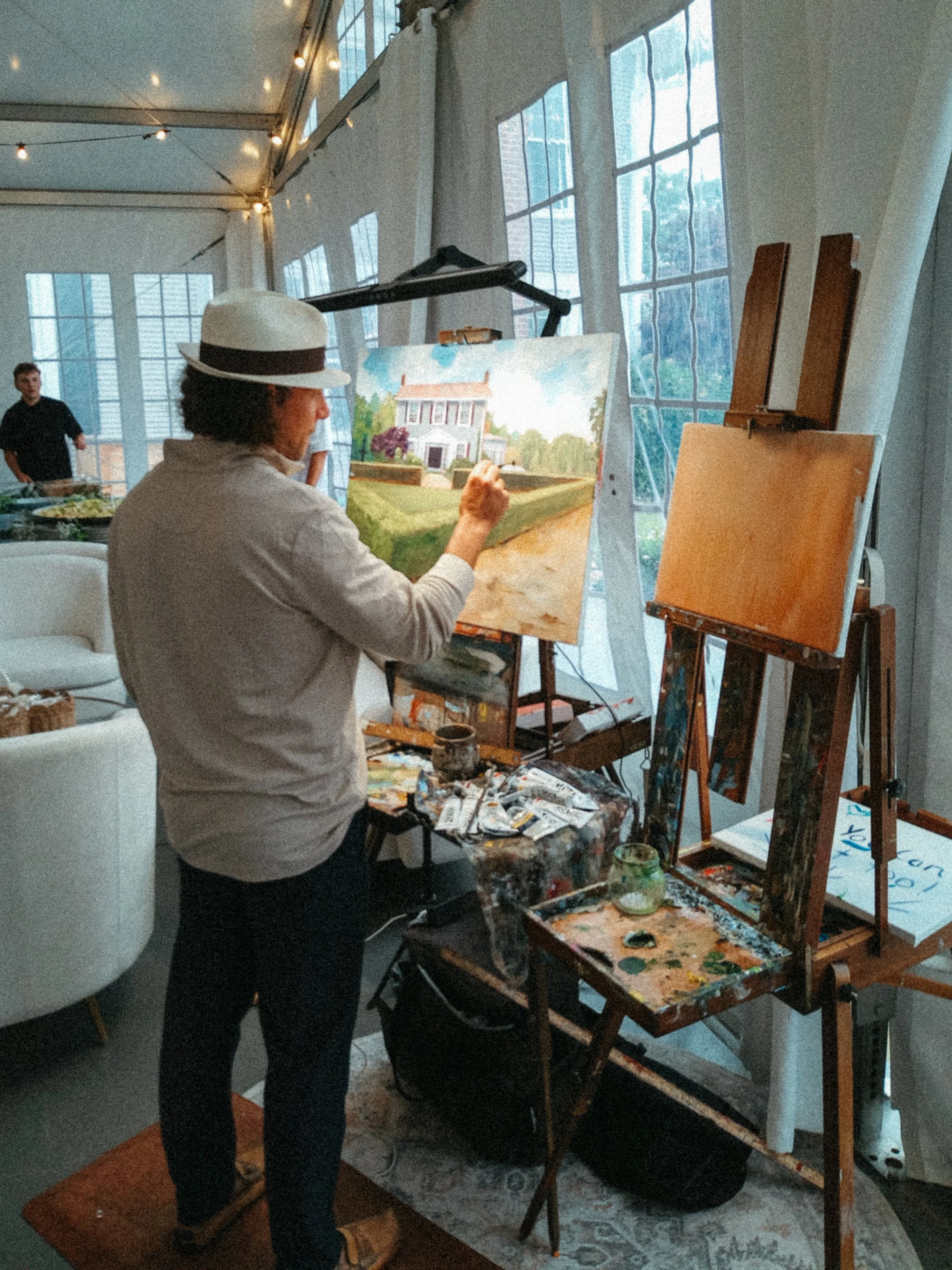 A woman wearing a white hat and white shirt is painting a landscape scene on a canvas at an art studio or gallery. She stands in front of an easel with her brush in hand, focusing on her work. The studio has large windows, natural light, and some other canvases, including an alt color canvas in the background. There are a few people in the background, and the room is decorated with string lights.