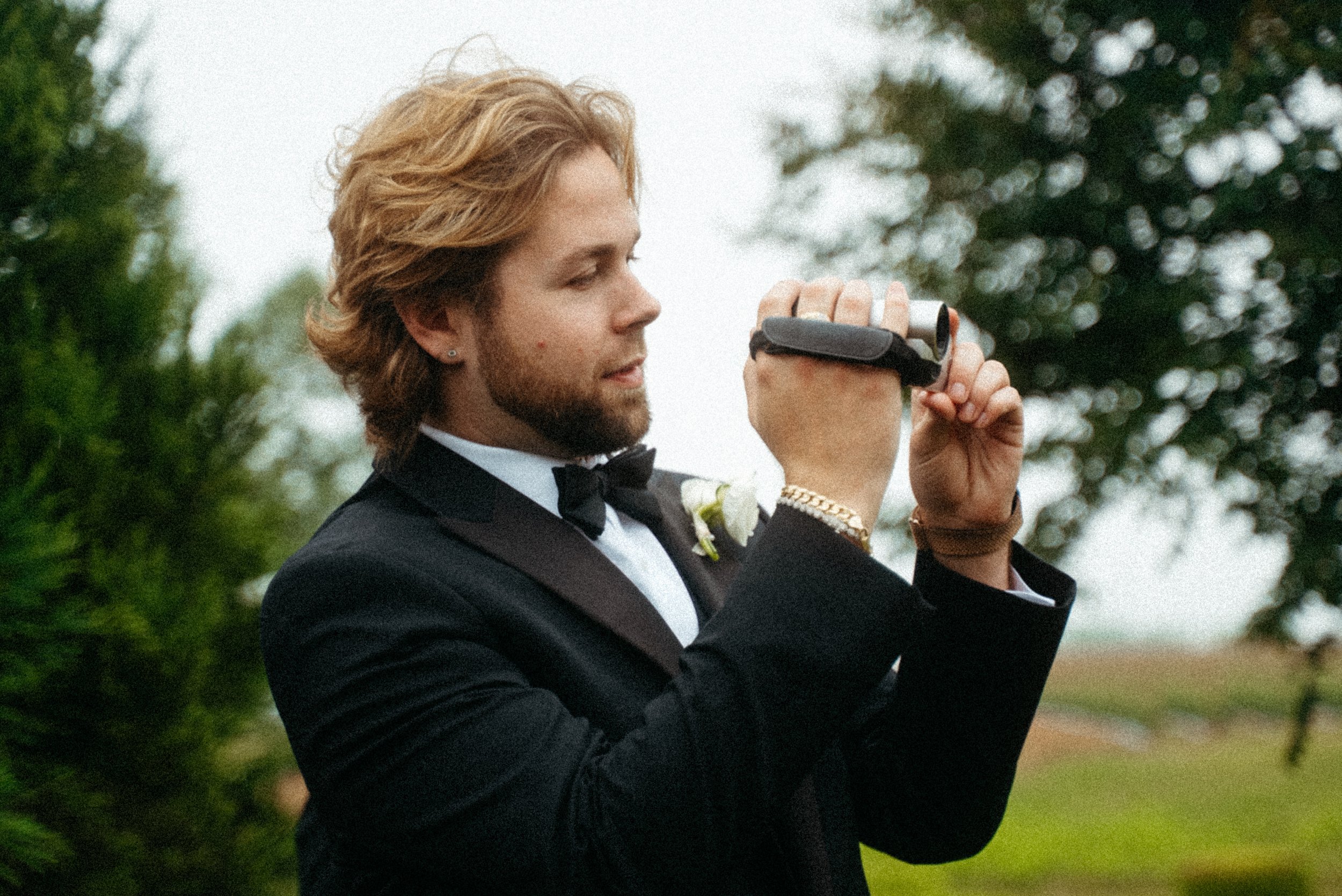 A man in a wedding suit with styled blond hair and a beard wearing a tuxedo and bow tie, holding a vintage camcorder to his eye outdoors with trees and a cloudy sky in the background.