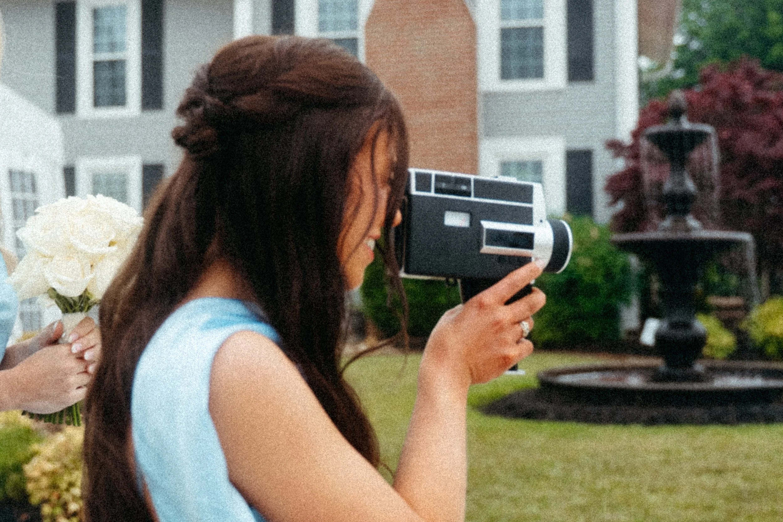 A woman in a bridesmaid dress with long brown hair taking a photo with a vintage super 8 style camera outdoors, with people embracing in the background and a residential building and greenery nearby.