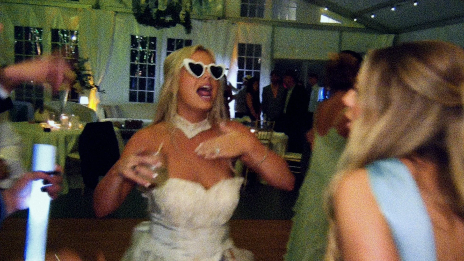 A woman in a wedding dress with heart-shaped sunglasses at a party or celebration. Taken on a disposable camera to give a nostalgic, vintage look.