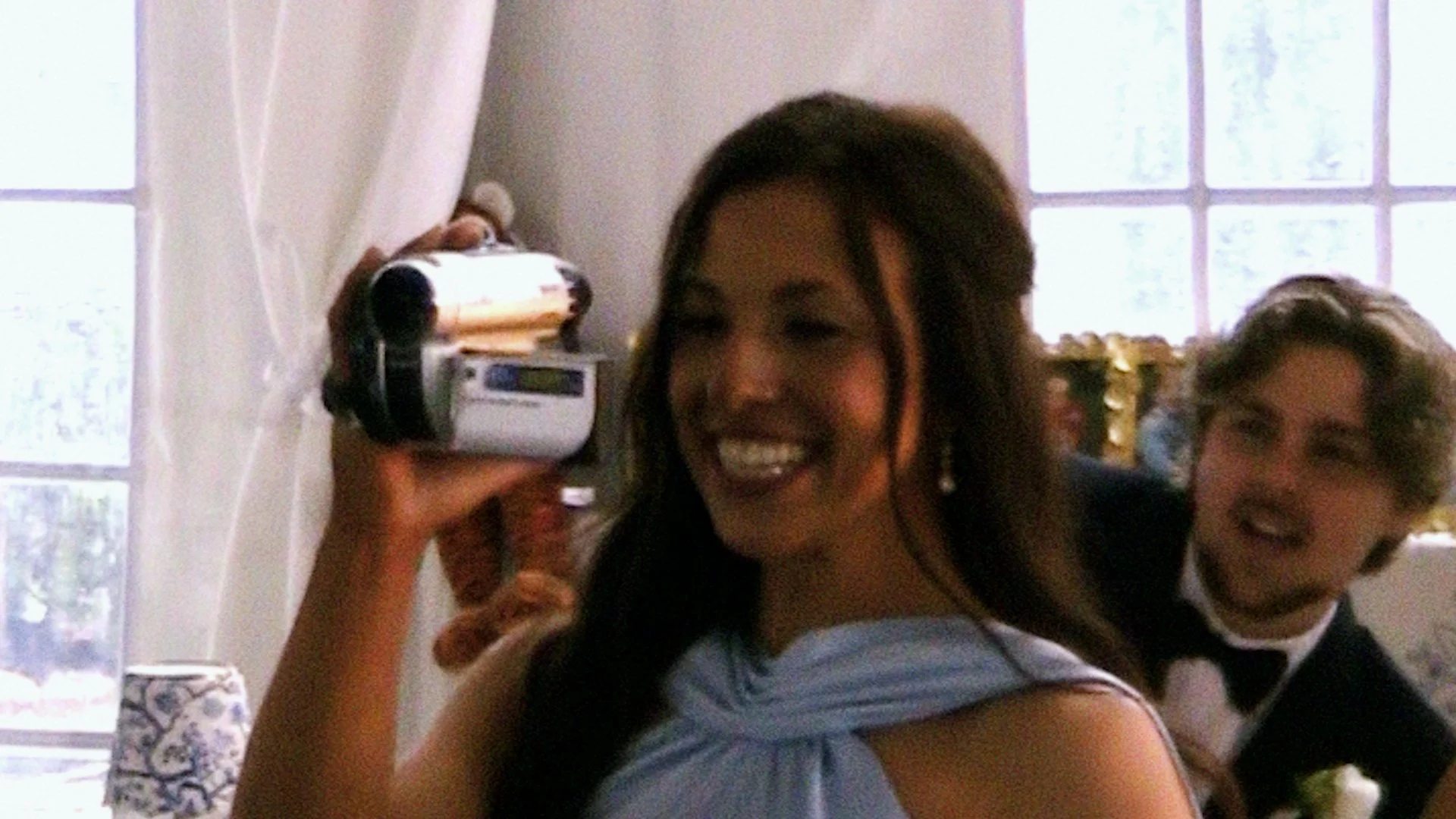 A young woman smiling as she films a wedding with a camcorder with a groom smiling beside her, in a well-lit room with large windows.