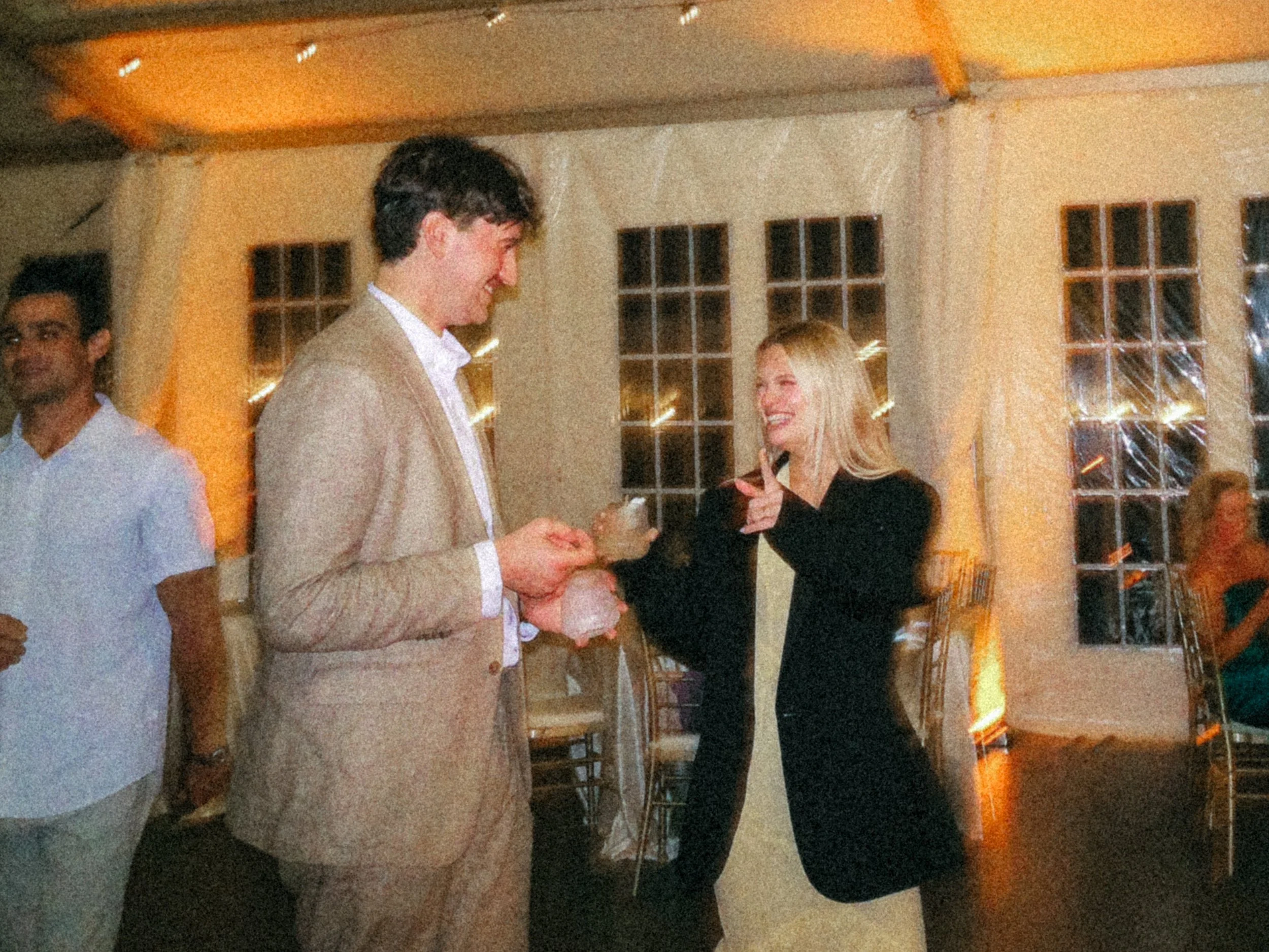 A group of people at a social gathering inside a room with large windows, warm lighting, and a wood ceiling. Two individuals are in focus, smiling and holding drinks, engaged in conversation. Taken on a disposable camera to give a nostalgic, vintage