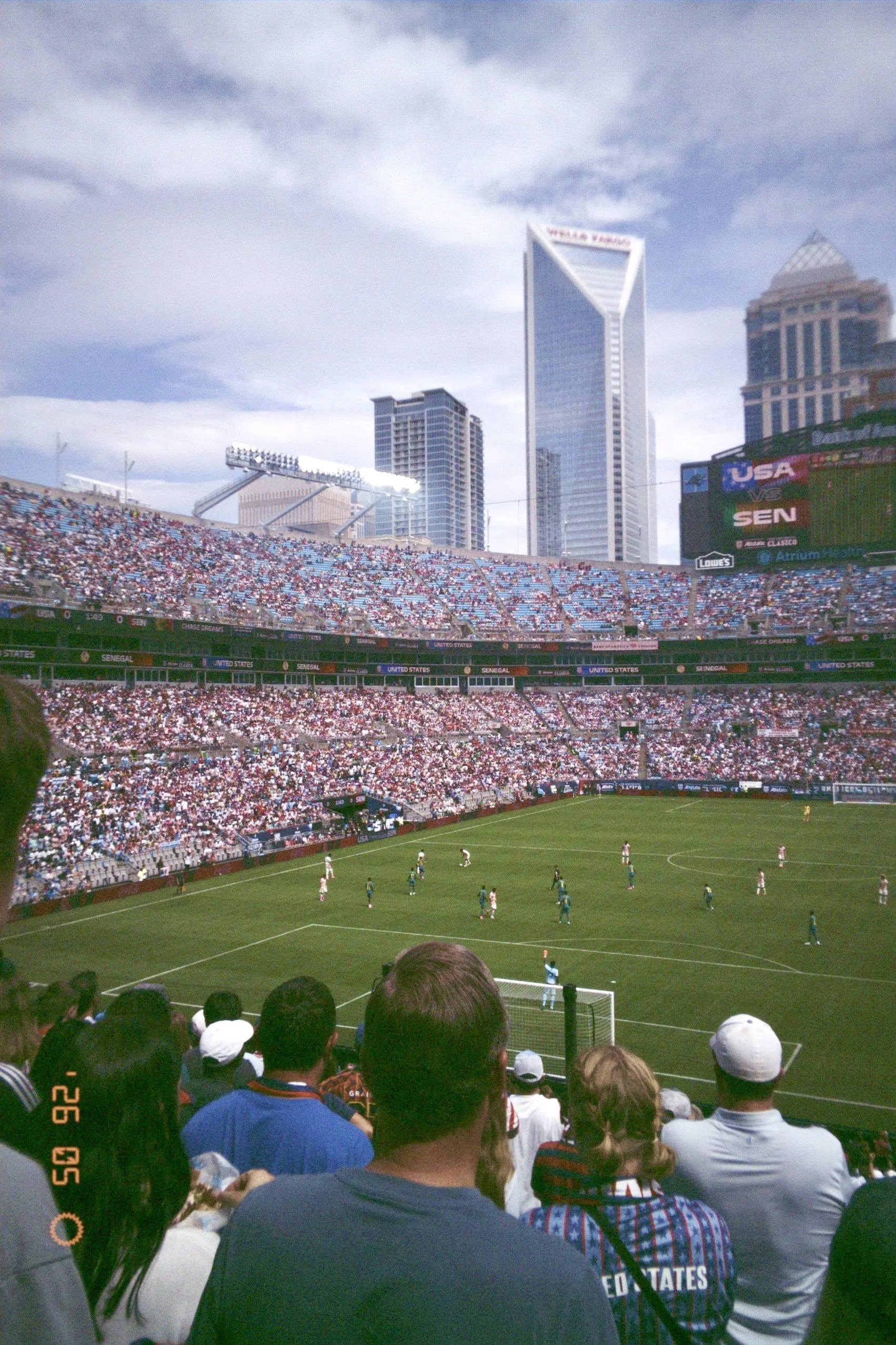 Crowd of spectators watching a soccer match at a stadium in an urban city, with tall skyscrapers in the background and a large digital scoreboard displaying USA and Senegal flags. Taken on a disposable camera to give a nostalgic, vintage look.