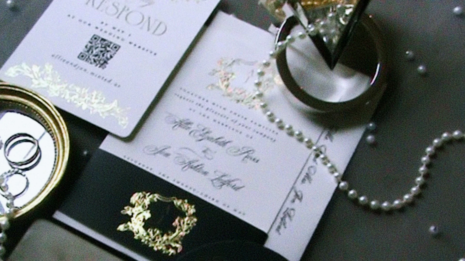 A wedding invitation card with elegant cursive text, gold wedding rings in an open ring box, a pearl necklace, a jar of candies, a decorative ribbon, and some scattered golden confetti on a black surface. Taken on a disposable camera