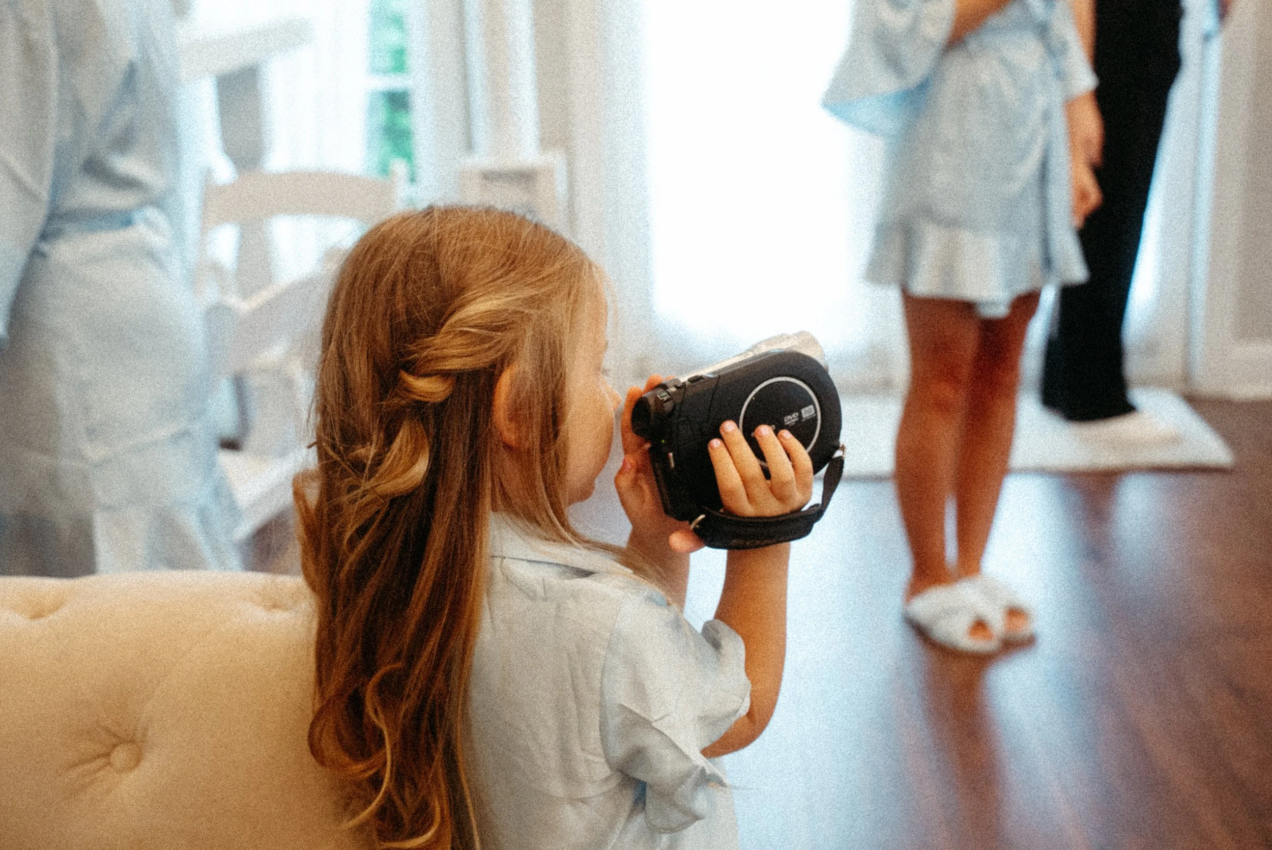A young girl with long red hair is sitting on a beige tufted chair, holding a black camera up to her eye and taking a photo. She is indoors, surrounded by people, some of whom are dressed in light-colored clothing.
